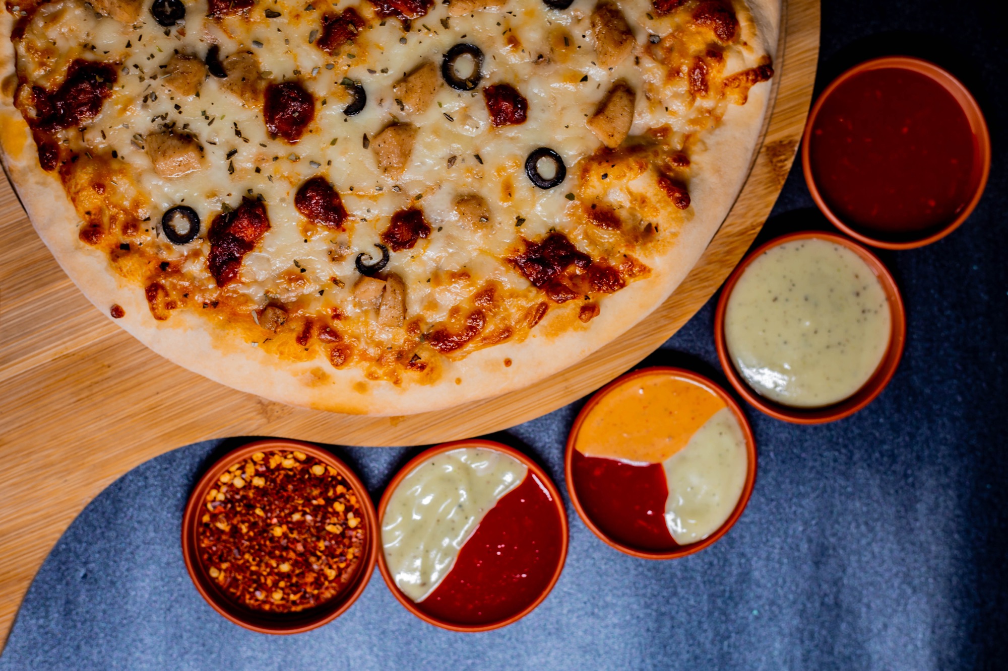 Overhead shot of a whole pizza with a trio of dipping sauces on a wooden board