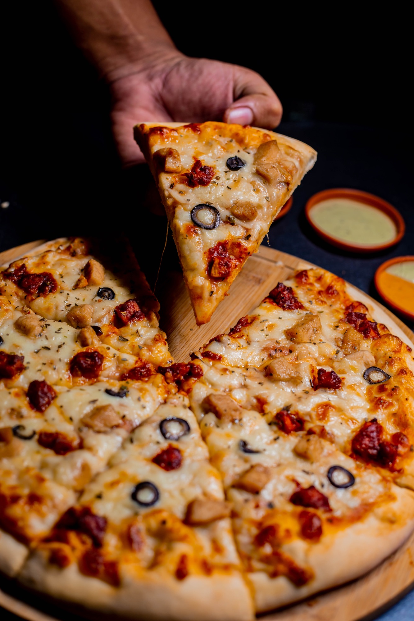 Hand lifting a slice from a freshly baked pizza with dipping sauce on the side
