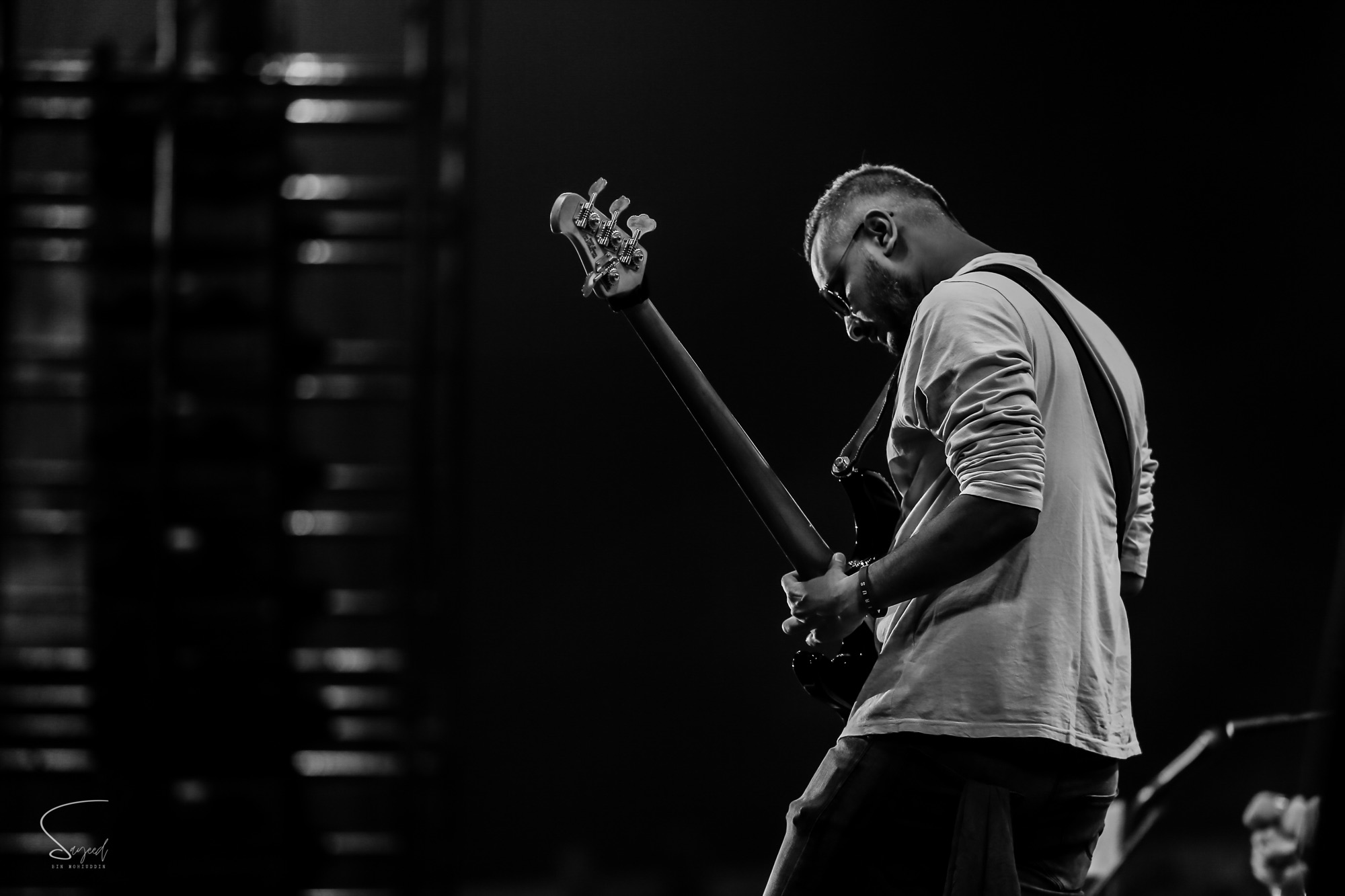 Bassist seen from behind, head bowed over his instrument in a moody black-and-white concert photo from Dhaka — Sayeed BM