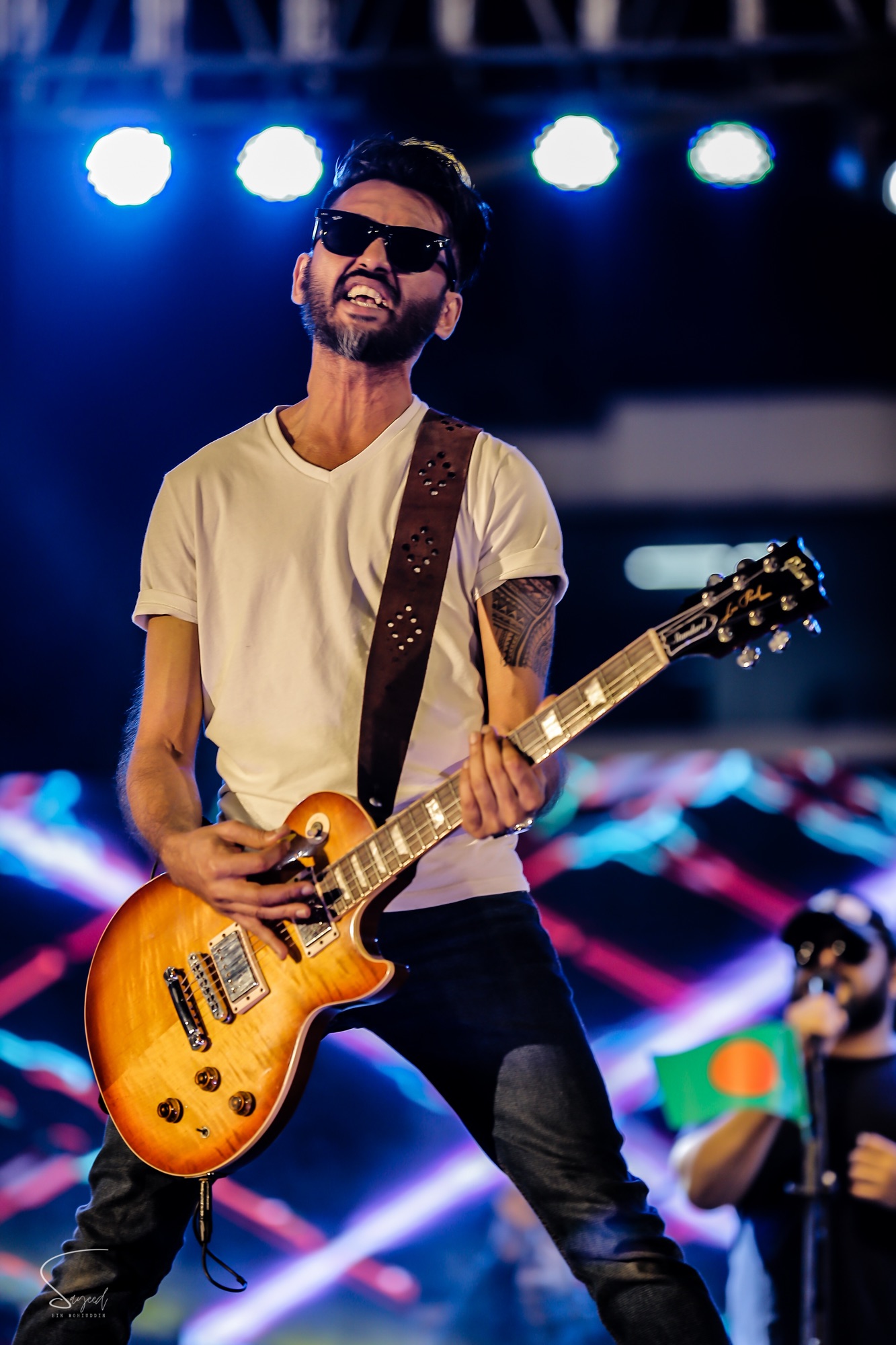 Guitarist in sunglasses belting it out with a sunburst guitar under blue stage lights at a Dhaka concert — Sayeed BM