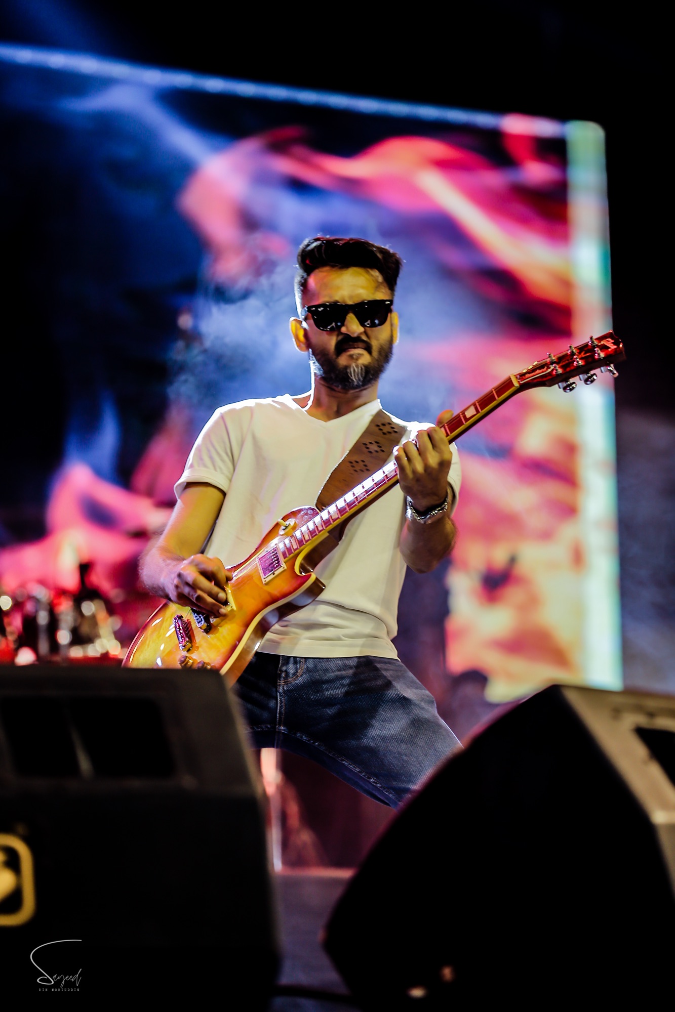Suharto Sheriff playing guitar in sunglasses against fiery red projected visuals at an Arbovirus concert in Dhaka — Sayeed BM