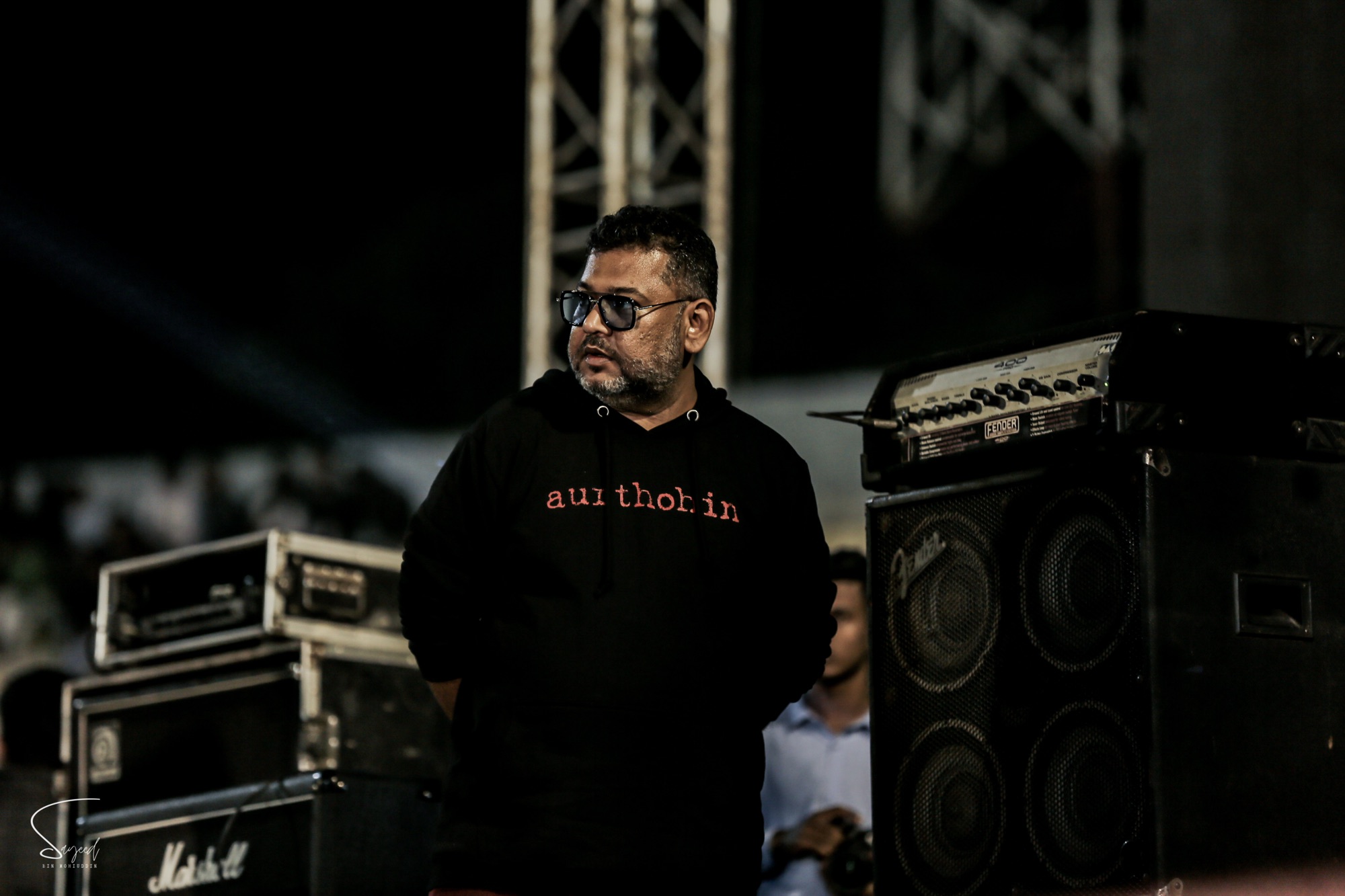 Man in an Aurthohin hoodie standing near amplifiers backstage at a Dhaka concert — Sayeed BM