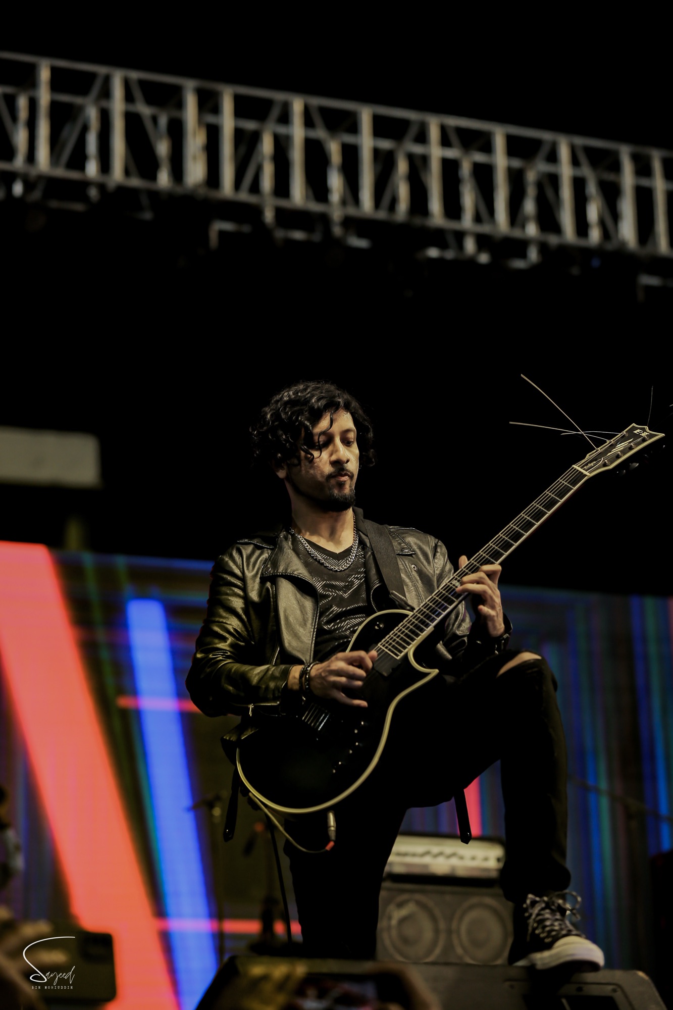 Guitarist in a leather jacket crouched on stage, foot raised, colorful stage lights at a Dhaka concert — Sayeed BM