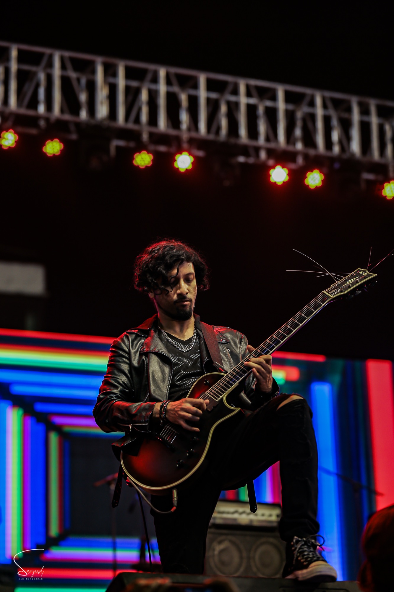 Guitarist in a leather jacket leaning into the mic, foot propped on a keyboard, black-and-white concert shot in Dhaka — Sayeed BM
