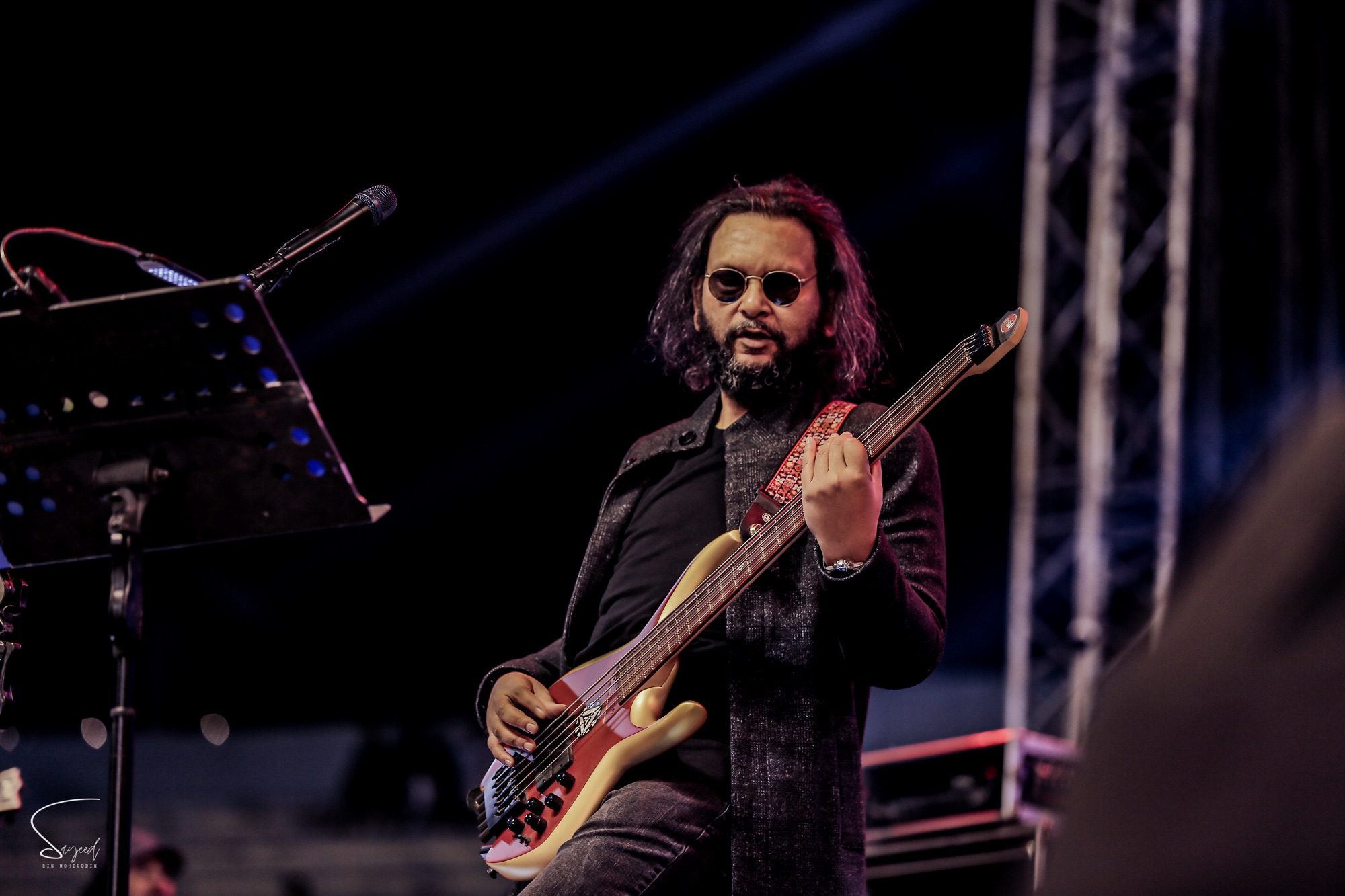 Bassist in round glasses playing a red five-string bass on stage at a Dhaka concert — Sayeed BM