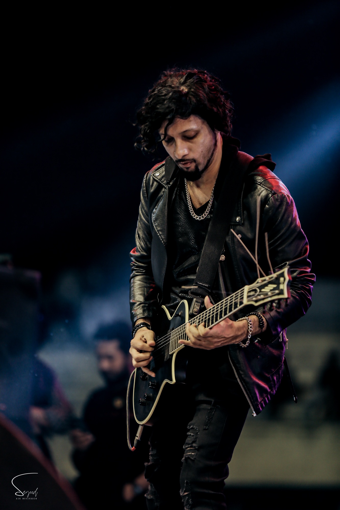 Guitarist with curly hair and leather jacket focused on his fretwork at a live concert in Dhaka — Sayeed BM