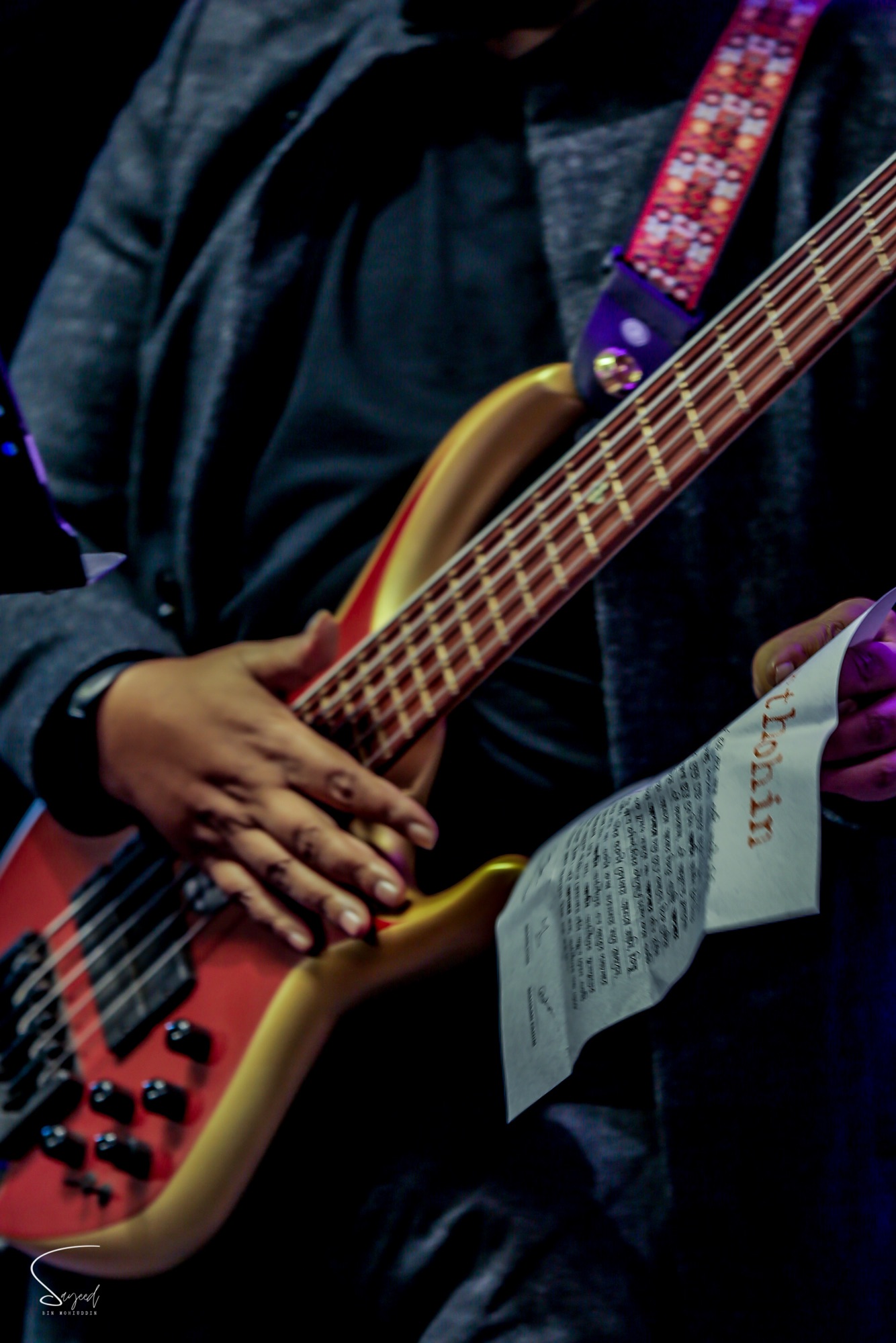 Close-up of hands fretting a red bass guitar while holding an Aurthohin setlist sheet at a Dhaka concert — Sayeed BM