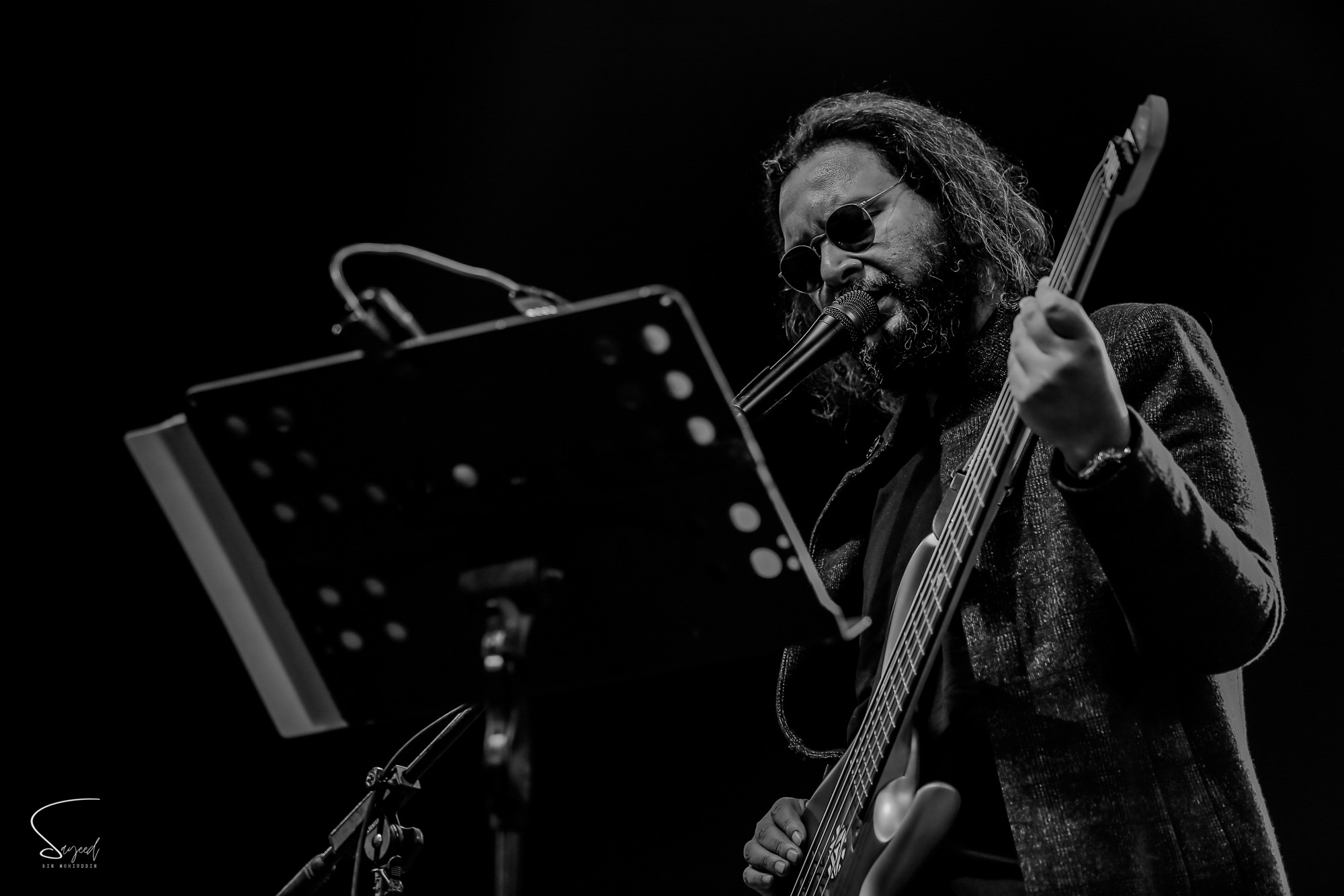Bearded bassist in sunglasses singing into the mic beside a music stand, black-and-white concert photo from Dhaka — Sayeed BM