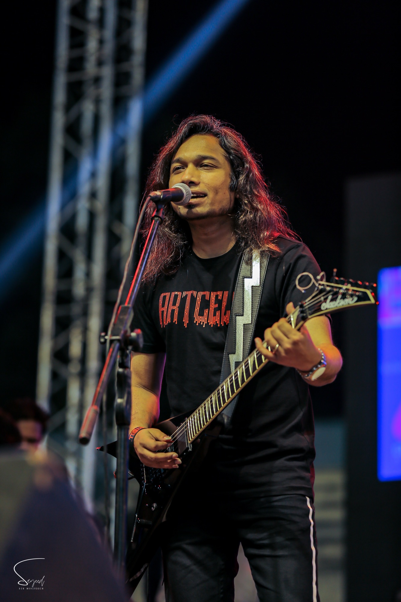 Guitarist in an Artcell t-shirt singing into the mic on stage at a live concert in Dhaka — Sayeed BM