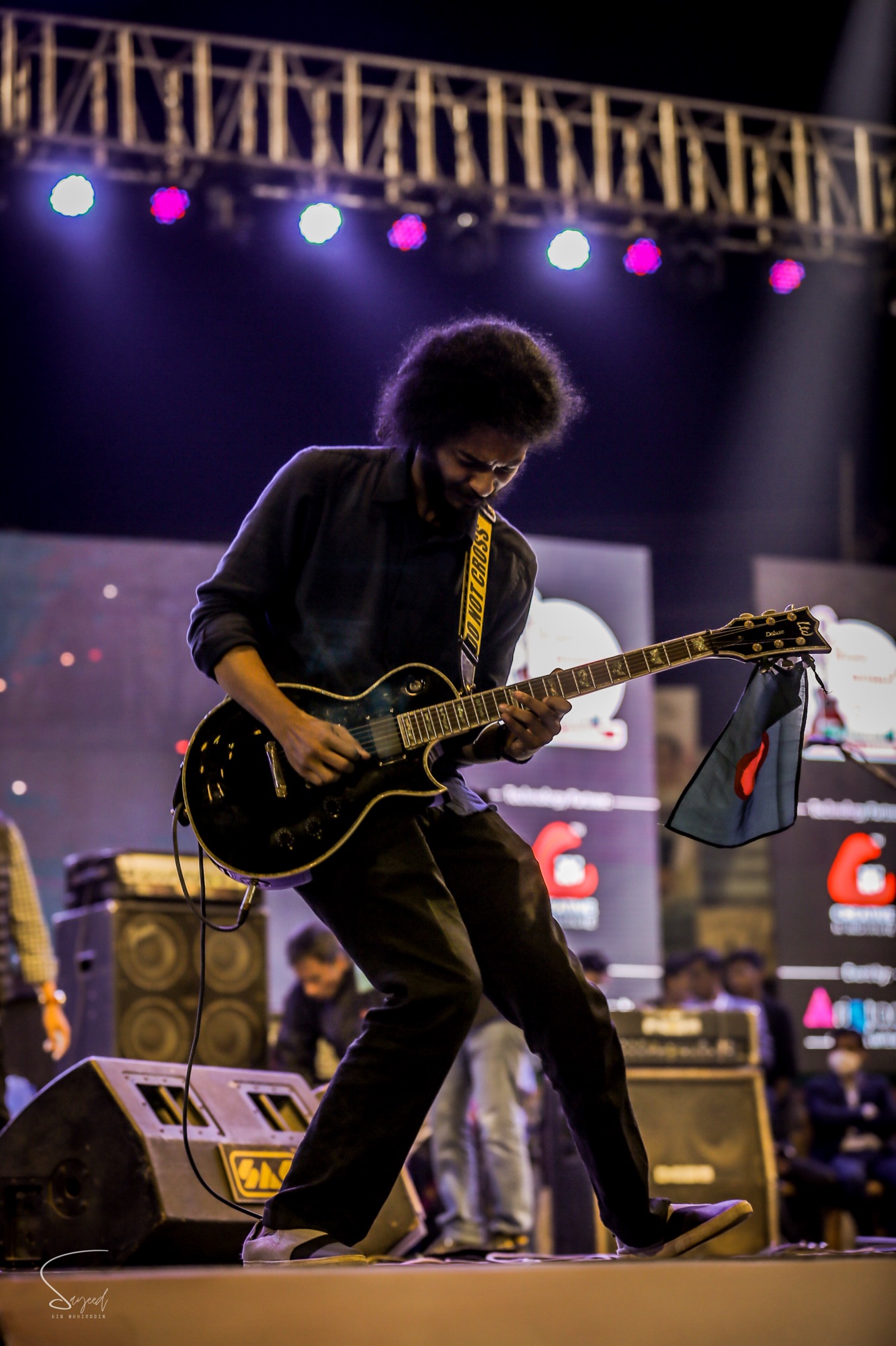 Guitarist with an afro lunging into a riff on stage, purple lights overhead at a concert in Dhaka — Sayeed BM