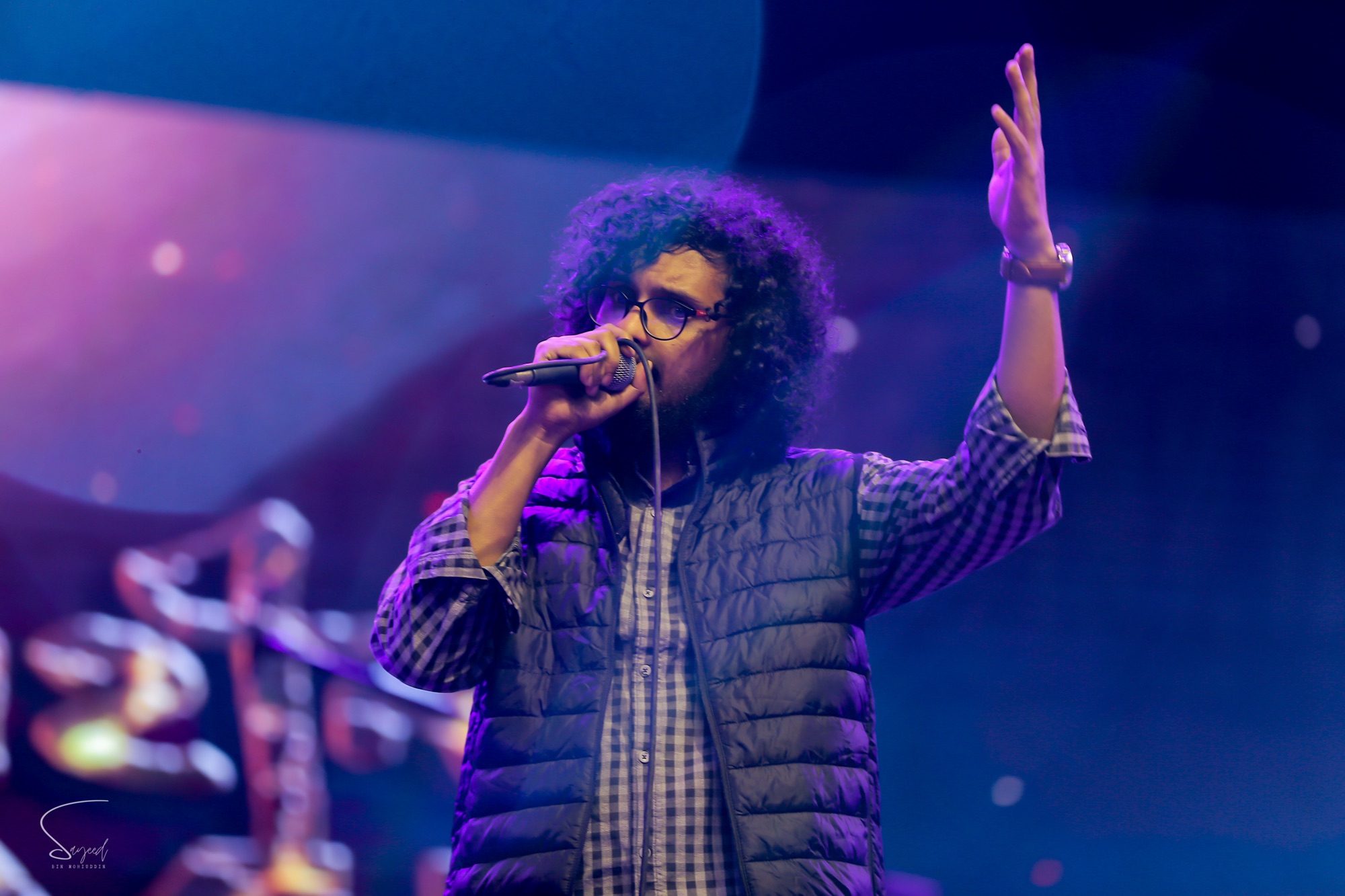 Vocalist with curly hair and glasses singing into a mic, hand raised under purple stage light at a Dhaka concert — Sayeed BM