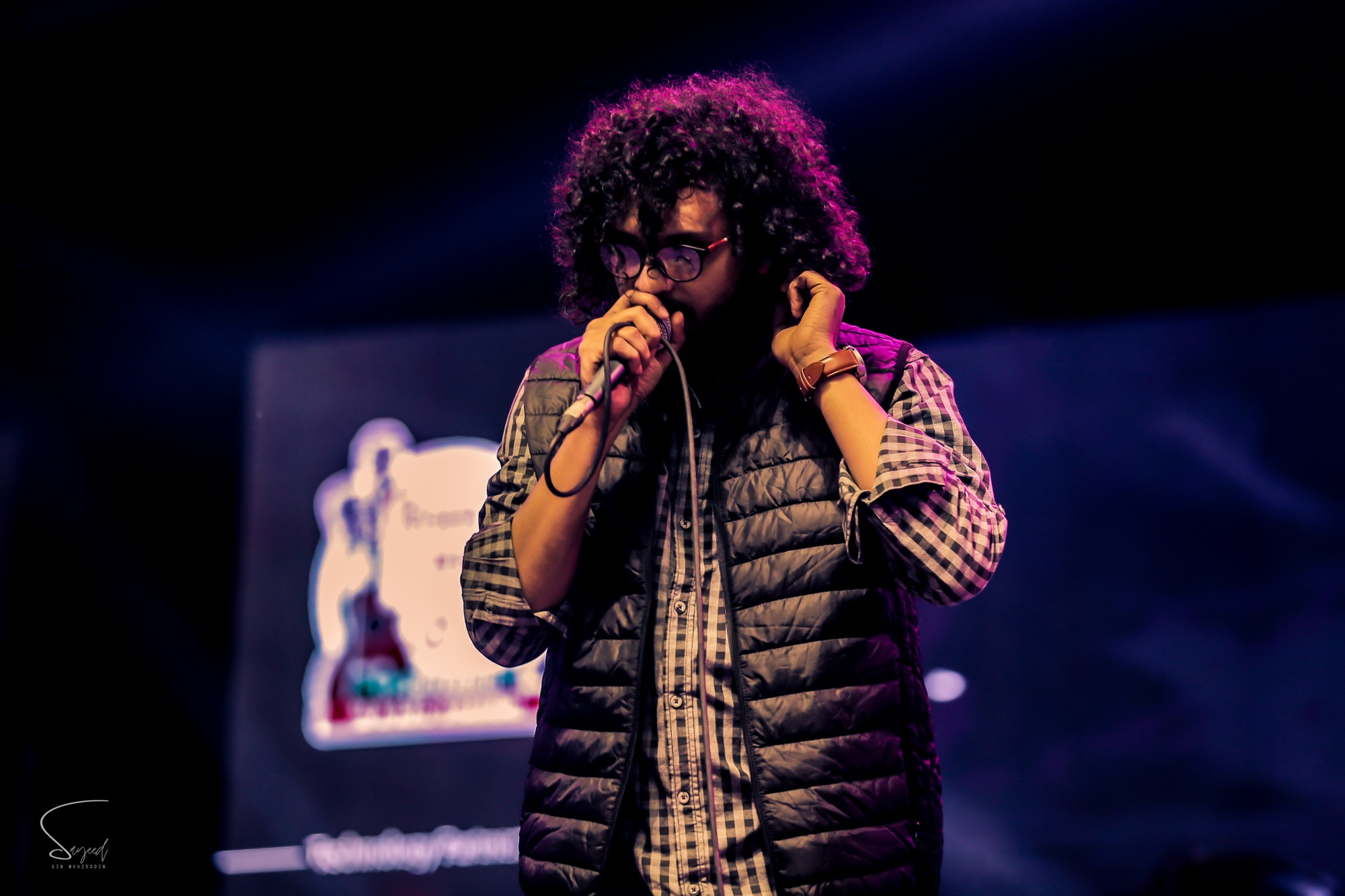 Singer with curly hair leaning into the microphone under magenta lighting at a live concert in Dhaka — Sayeed BM
