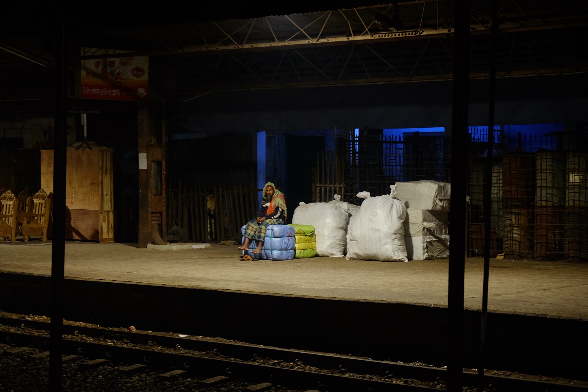 A traveller waits alone on a dark railway platform, single overhead light, Dhaka