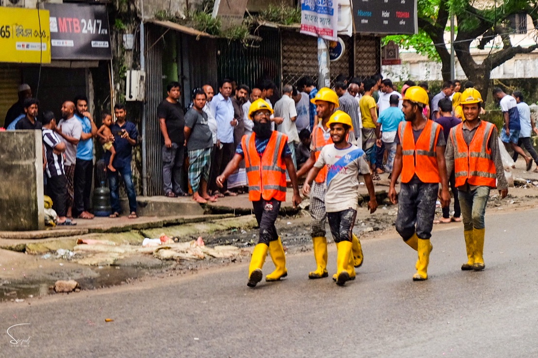 Construction workers in orange safety vests and helmets crowd a Dhaka street