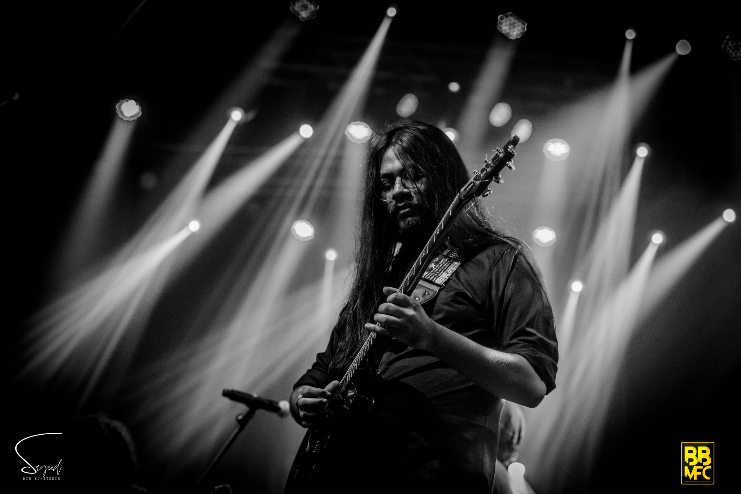 Long-haired guitarist framed by sweeping white light beams, black-and-white wide shot from a Dhaka concert — Sayeed BM