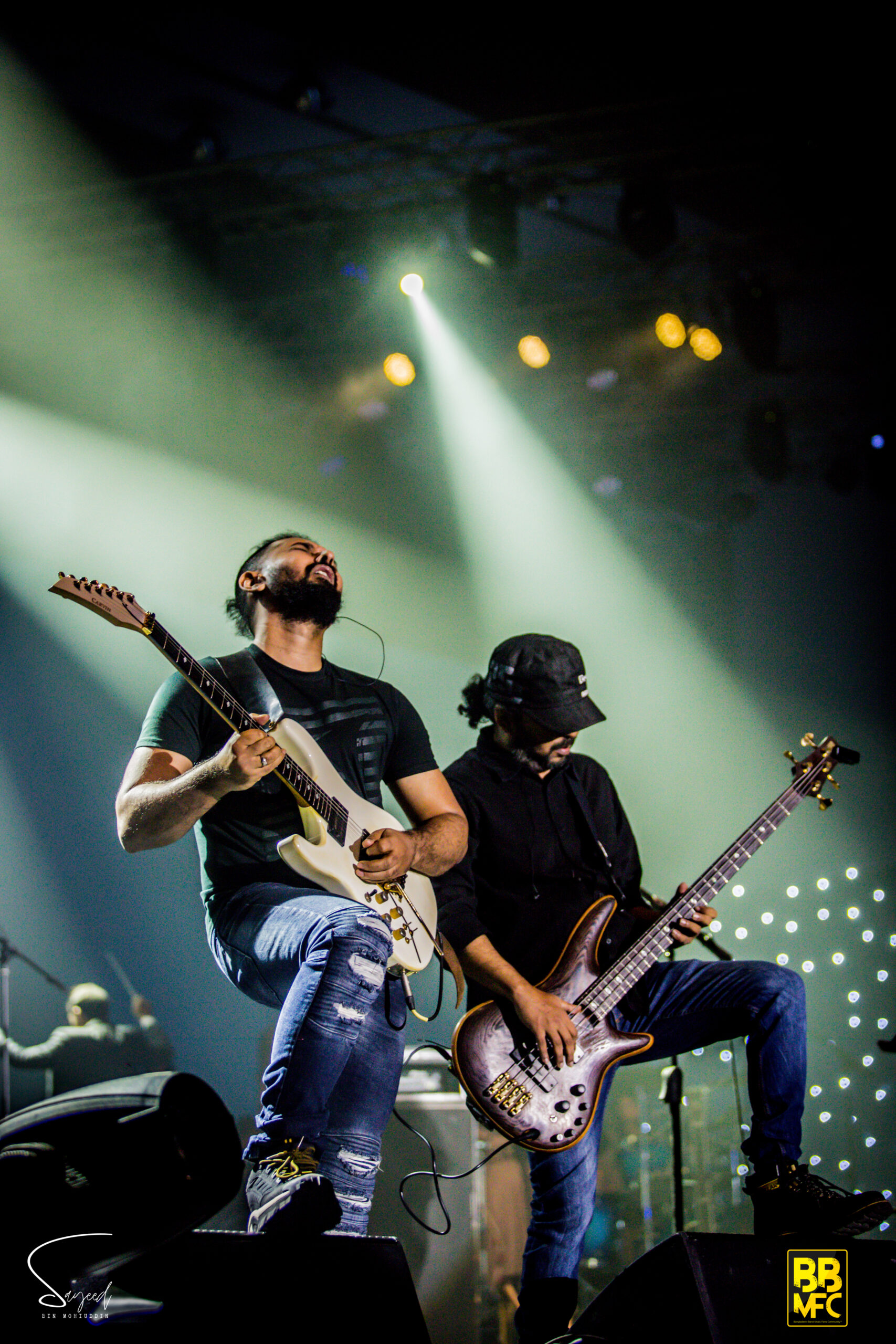 Guitarist leaning back mid-riff beside a bassist in a cap, lit by sweeping white stage beams in Dhaka — Sayeed BM