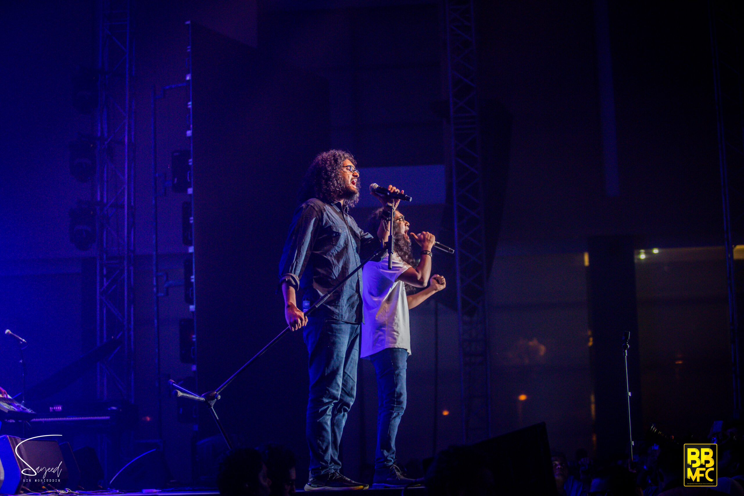 Two vocalists singing together into separate mics under deep blue stage lighting at a Shironamhin concert in Dhaka — Sayeed BM