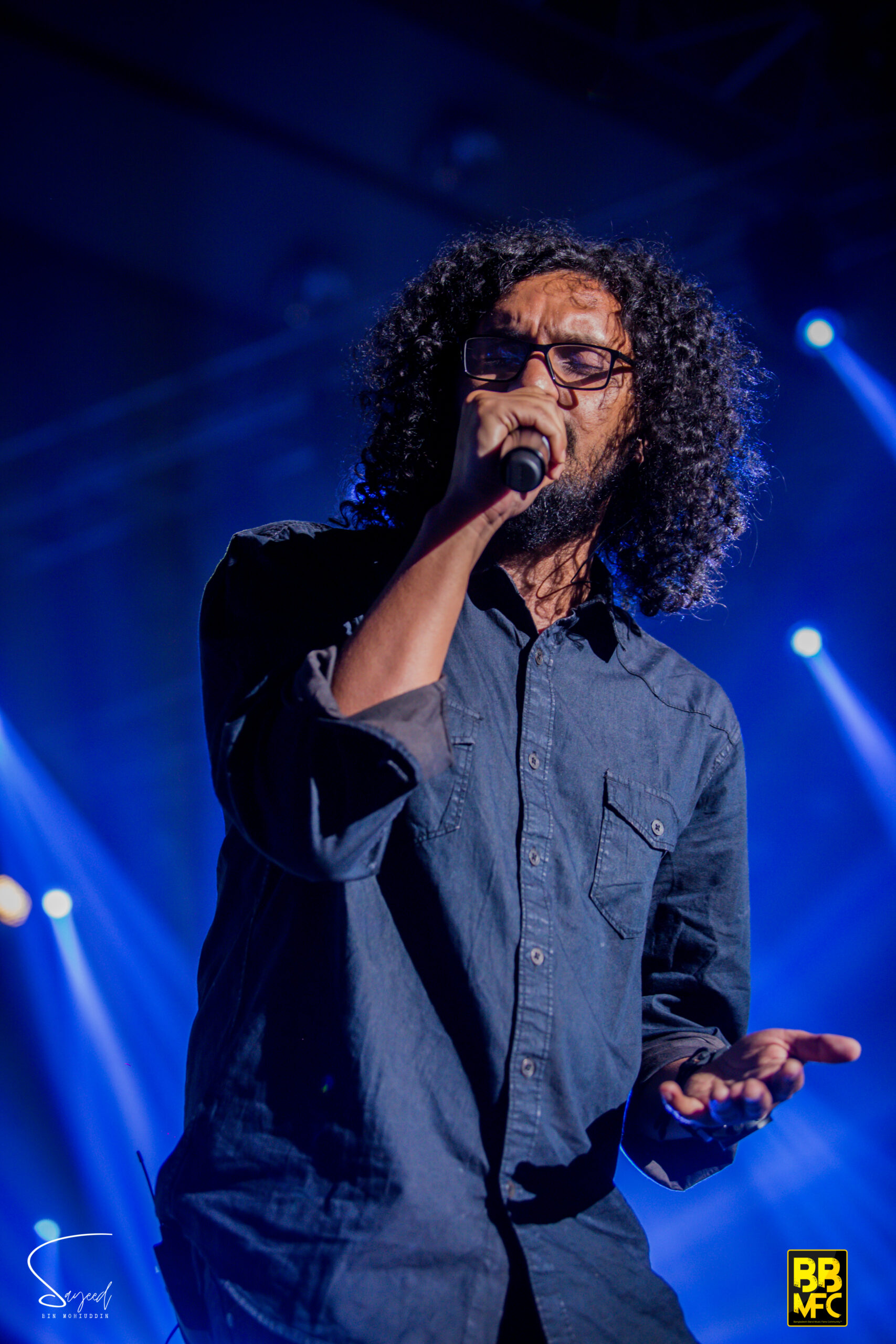 Curly-haired vocalist singing into the mic, bathed in deep blue stage light at a Shironamhin concert in Dhaka — Sayeed BM