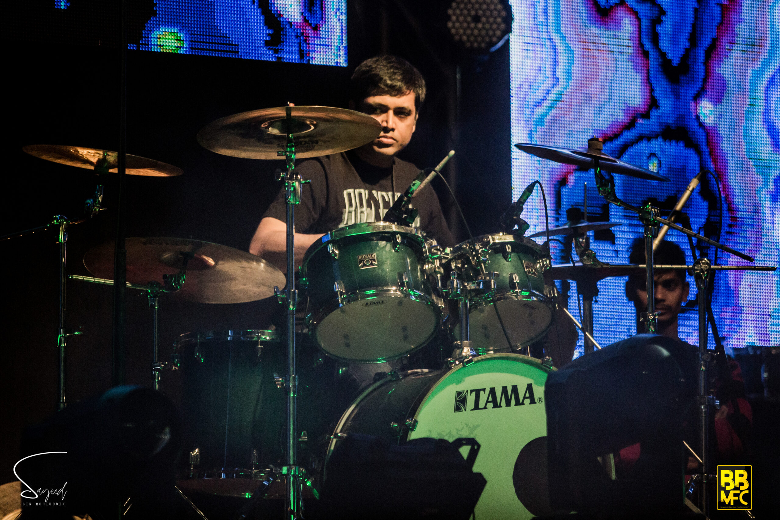 Drummer focused behind a green Tama drum kit, colorful LED visuals glowing behind him at a Dhaka concert — Sayeed BM