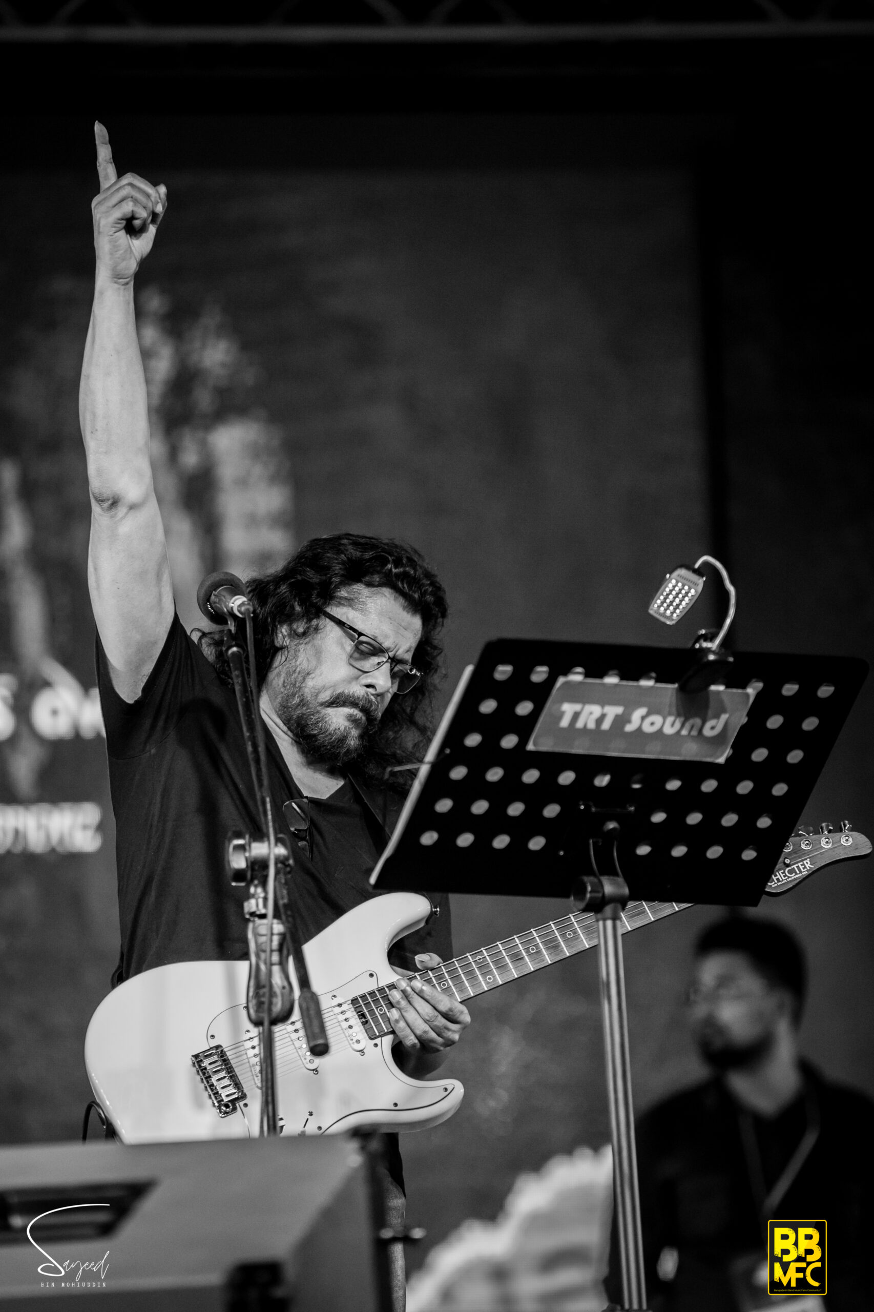 Guitarist raising a fist in the air mid-performance beside a sheet music stand, black-and-white concert photo from Dhaka — Sayeed BM