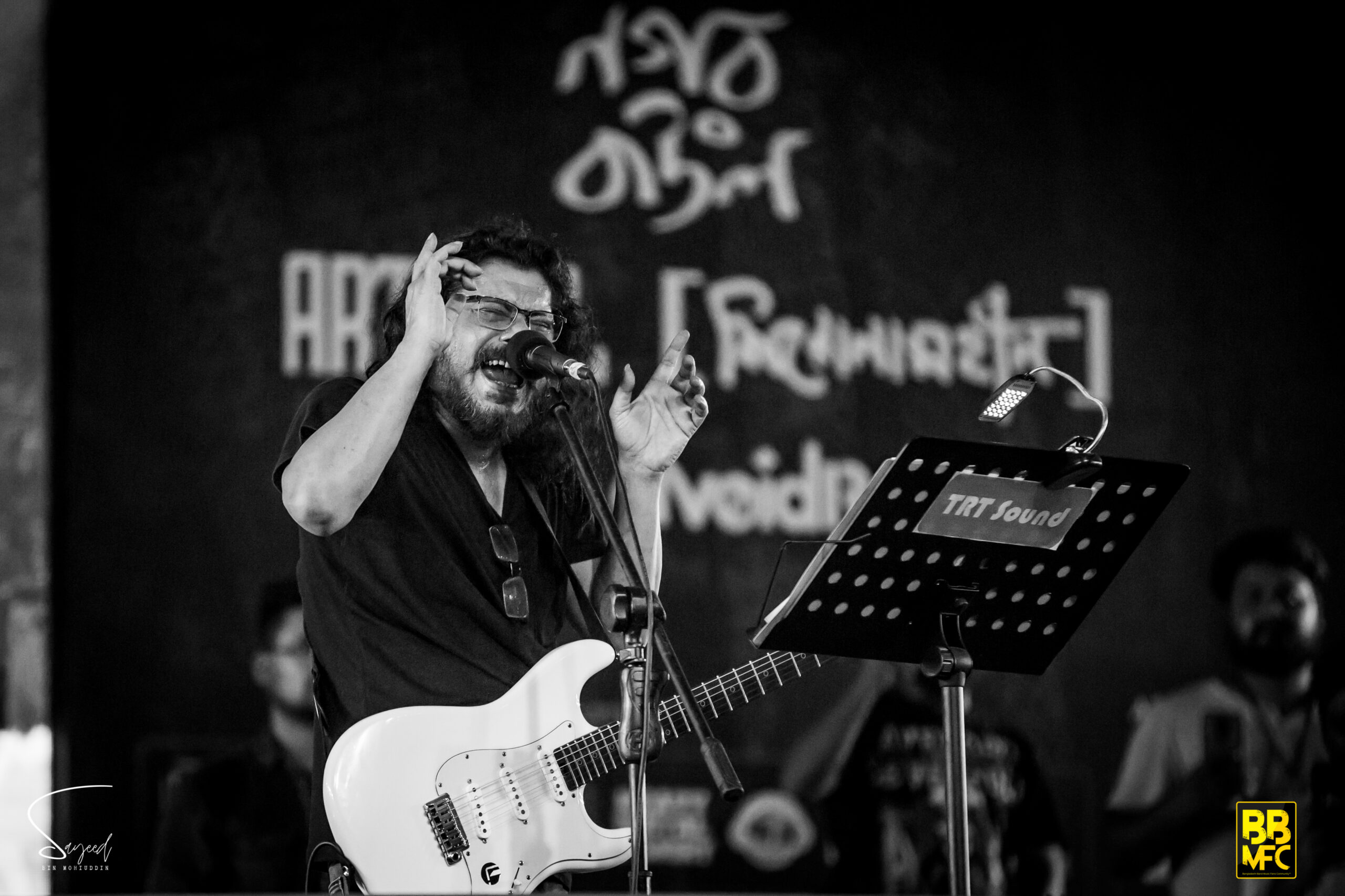 Singer with hands raised mid-performance beside a white guitar, black-and-white concert photo in Dhaka — Sayeed BM