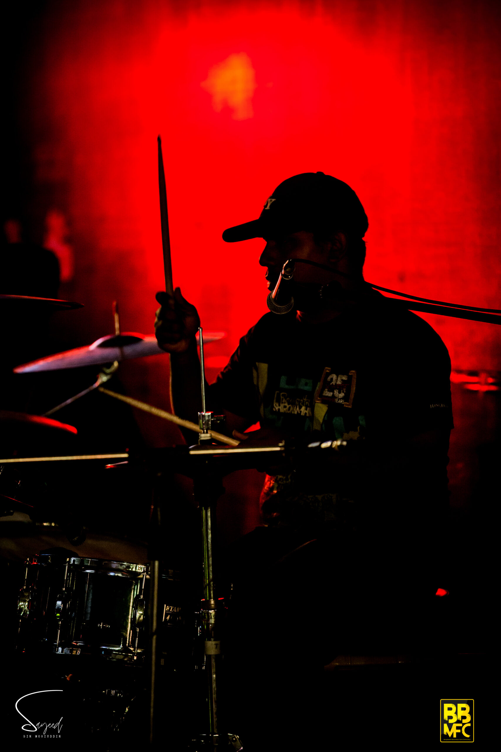 Drummer silhouetted against a blazing red backdrop, drumstick raised mid-strike at a Dhaka concert — Sayeed BM