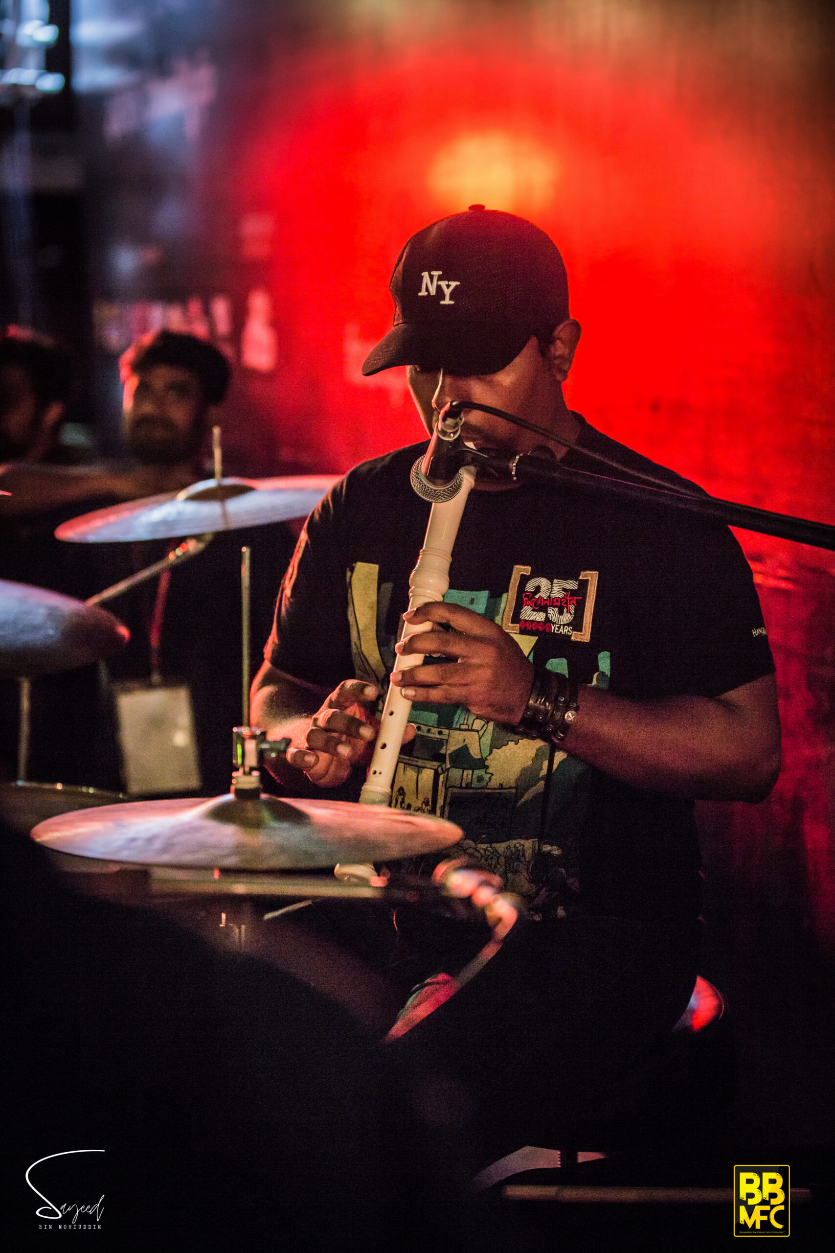 Drummer playing a flute one-handed beside his cymbals, lit by deep red stage lighting in Dhaka — Sayeed BM