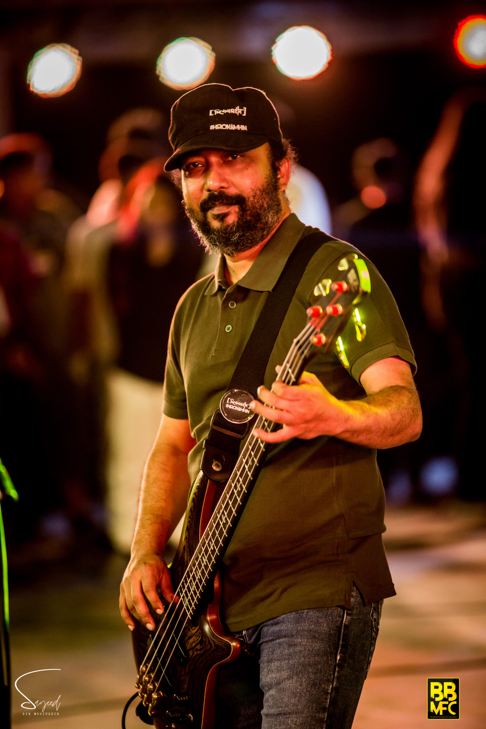 Bassist in a Shironamhin cap glancing toward the camera while performing in Dhaka — Sayeed BM