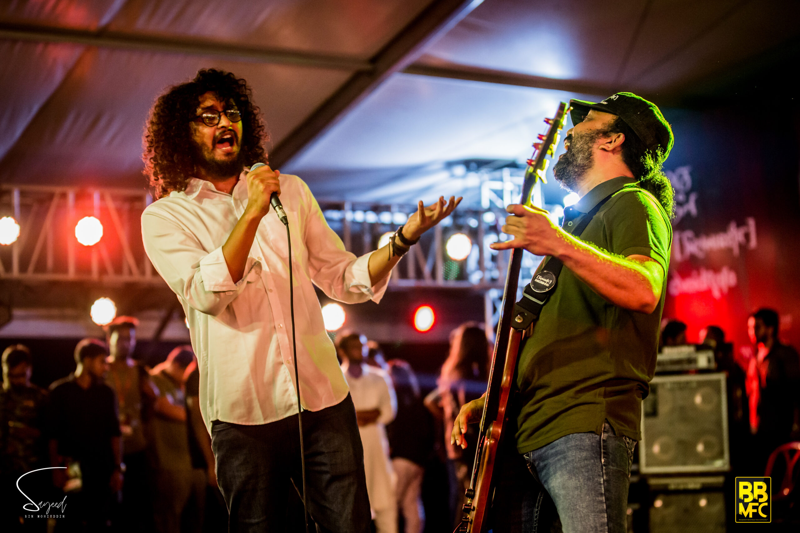 Two musicians sharing a mic mid-song, one playing guitar, lit by red and yellow stage lights at a Shironamhin show in Dhaka — Sayeed BM