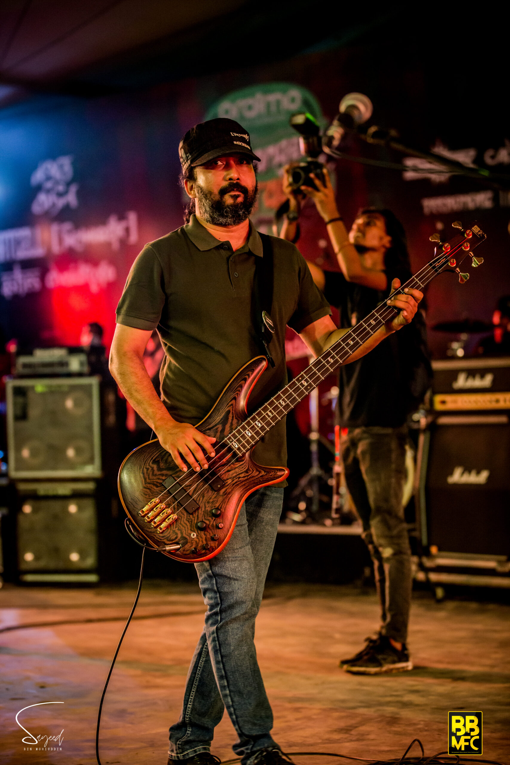 Bassist in a cap performing on stage as a bandmate raises a camera in the background at a Shironamhin concert in Dhaka — Sayeed BM