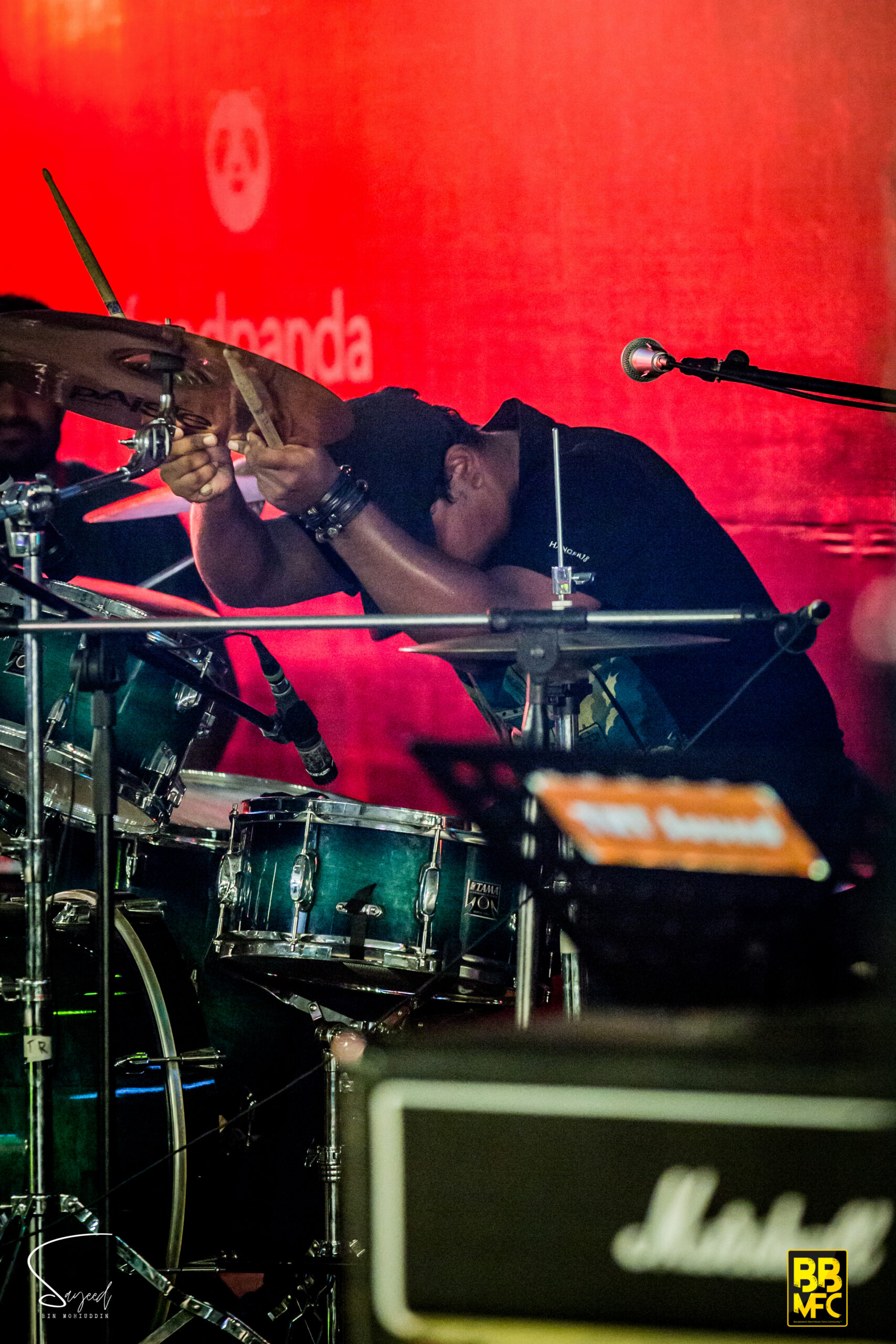 Drummer mid-strike on cymbals against a glowing red backdrop at a concert in Dhaka — Sayeed BM