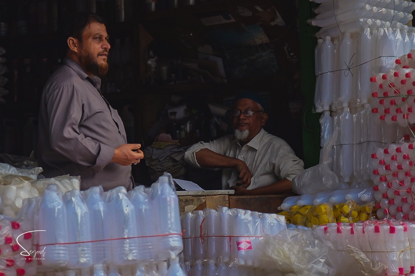 A night market vendor with stacked plastic bottles looks toward the camera, Dhaka