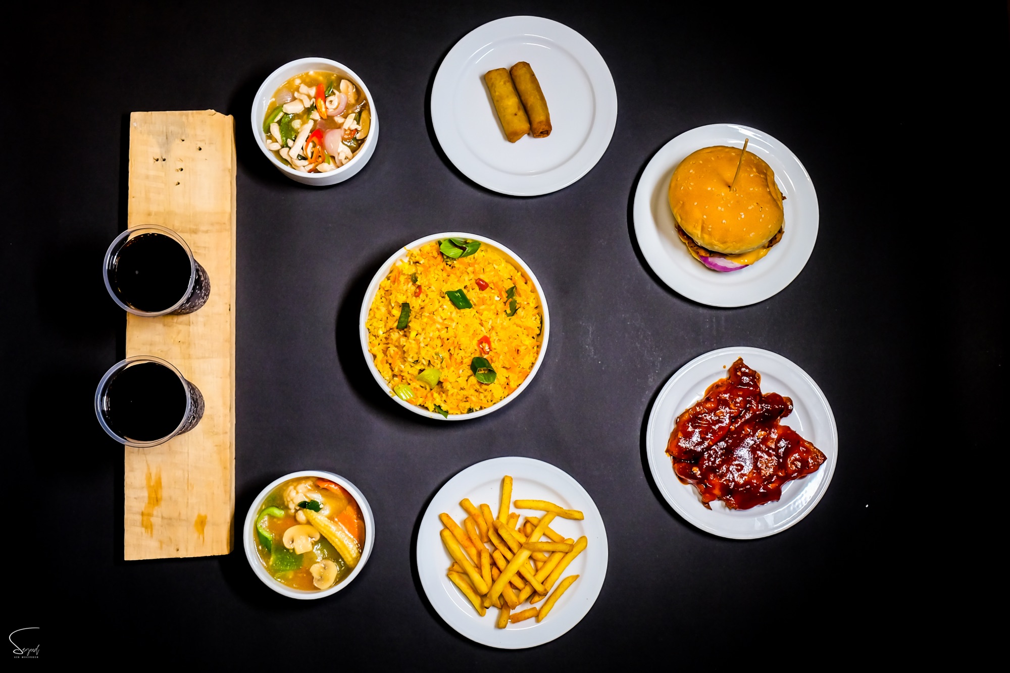 Overhead flat lay of curry, burger, fries and chutney bowls arranged on a dark backdrop