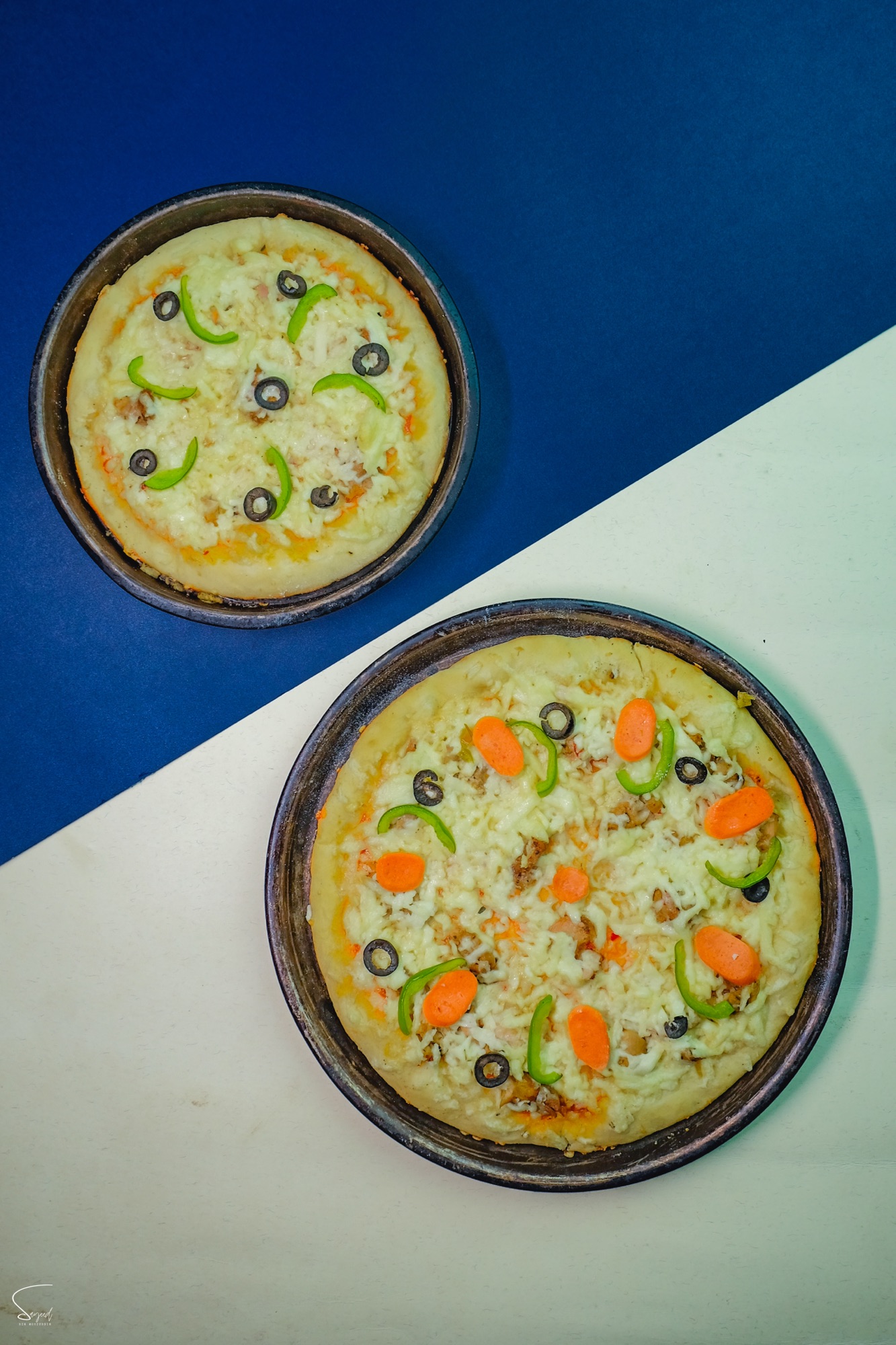 Two personal-size pizzas with olives and peppers shot overhead on a split blue and white background