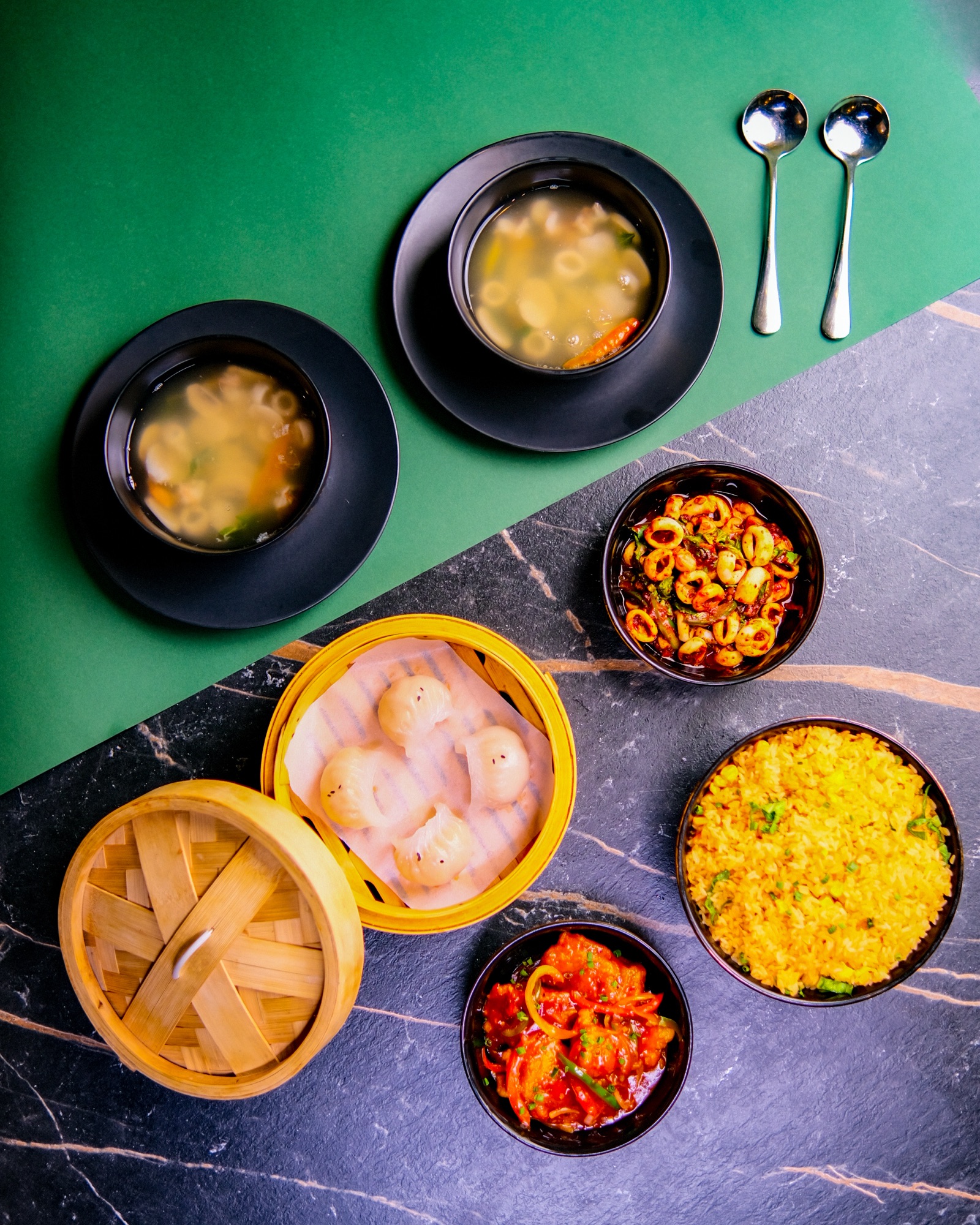 Overhead spread of dim sum bowls and soup with spoons on a green background
