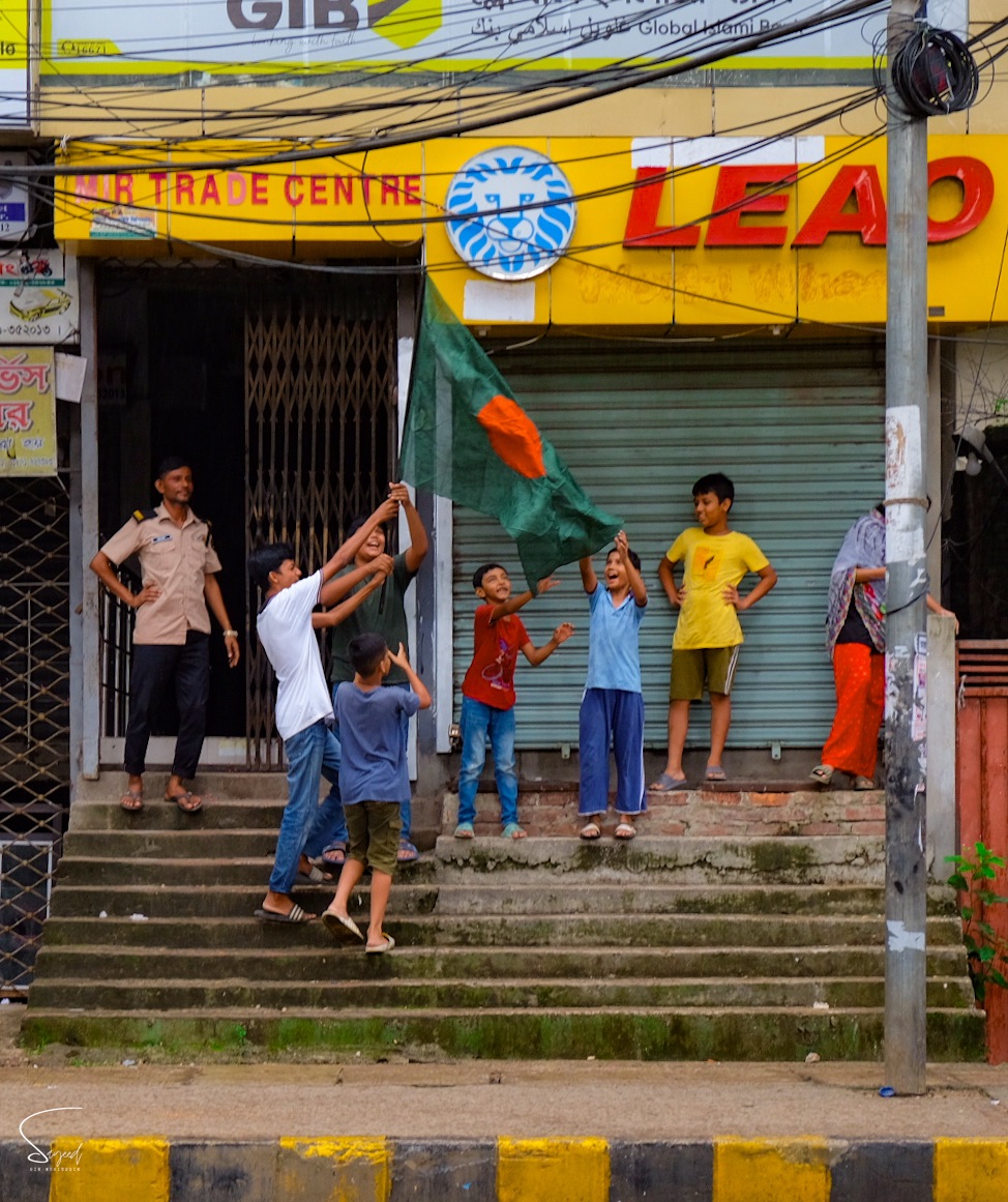Children waving Bangladesh flags in front of a colourful shop on Victory Day, Dhaka