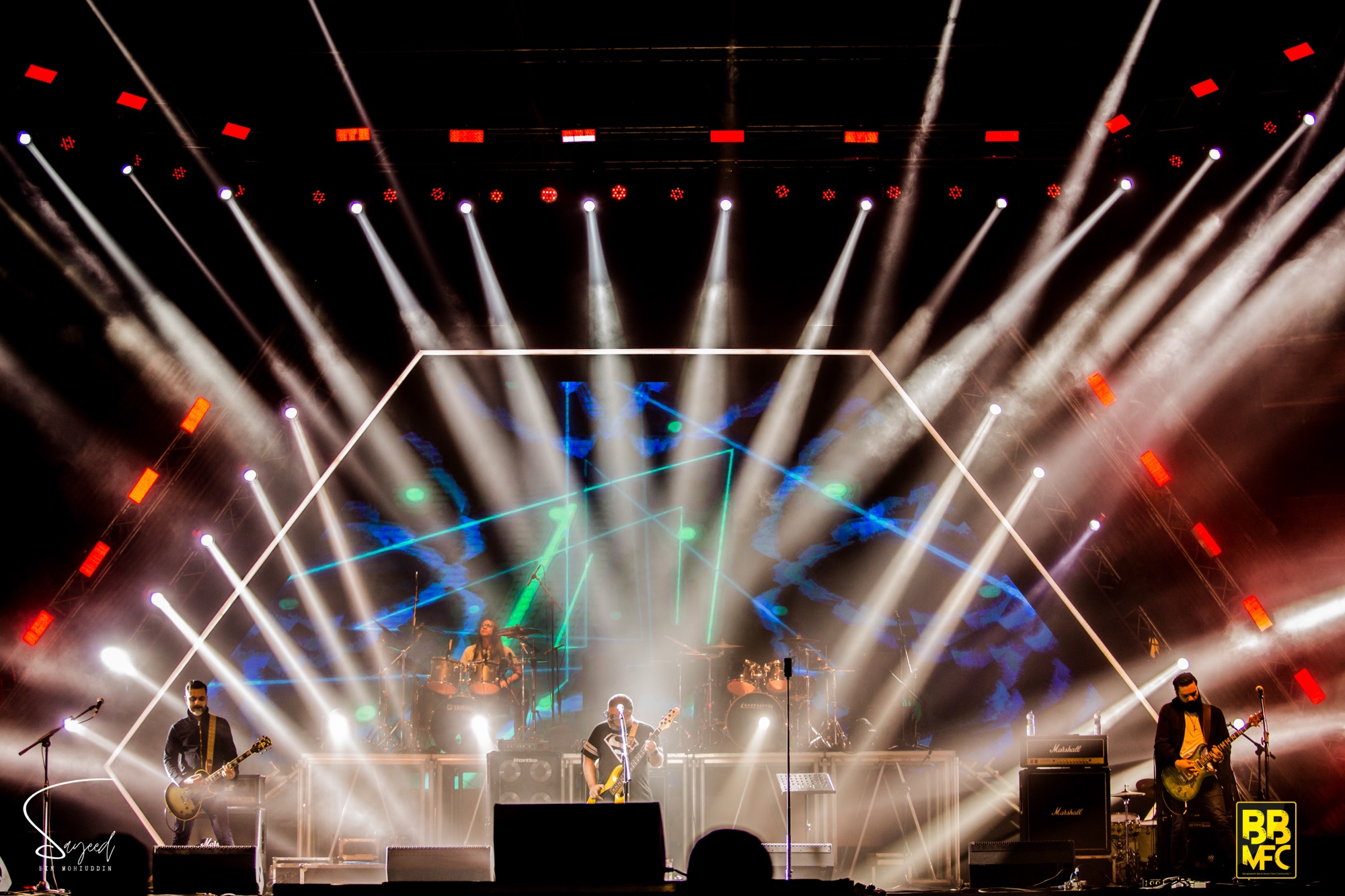 Wide shot of a full band on stage framed by a glowing hexagon light rig and fanning white beams at a live concert in Dhaka — Sayeed BM