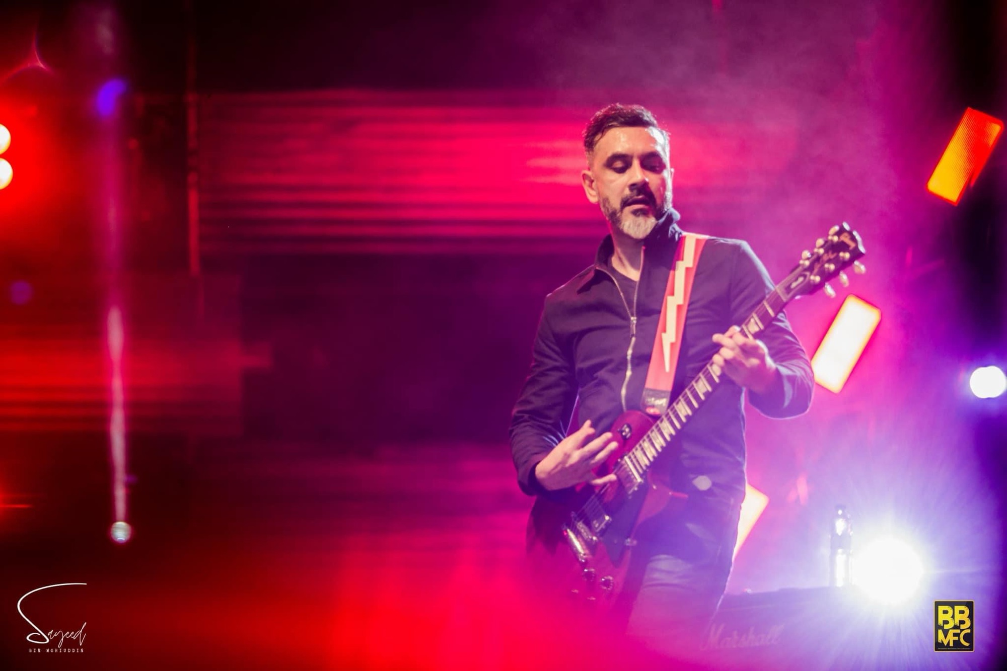 Guitarist with a grey beard playing a red guitar under red and purple stage light at a Shironamhin concert in Dhaka — Sayeed BM
