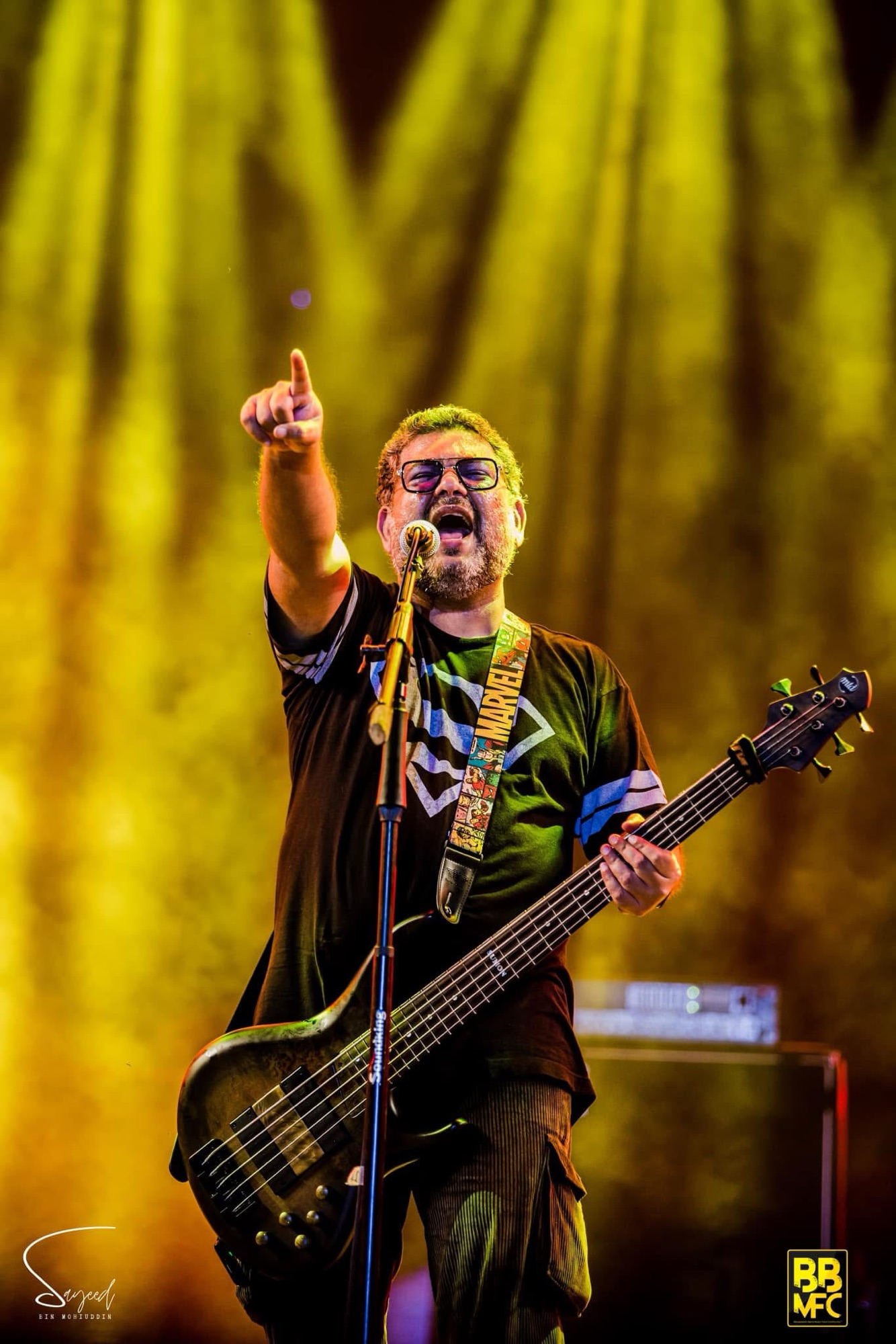 Bassist pointing skyward under golden stage light rays, vertical shot from a Shironamhin concert in Dhaka — Sayeed BM