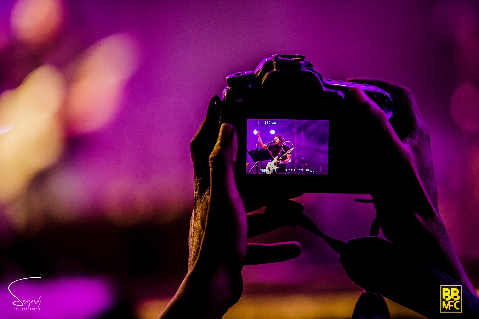 Camera viewfinder capturing a guitarist on stage, photographer's hands in frame under purple bokeh at a Dhaka concert — Sayeed BM