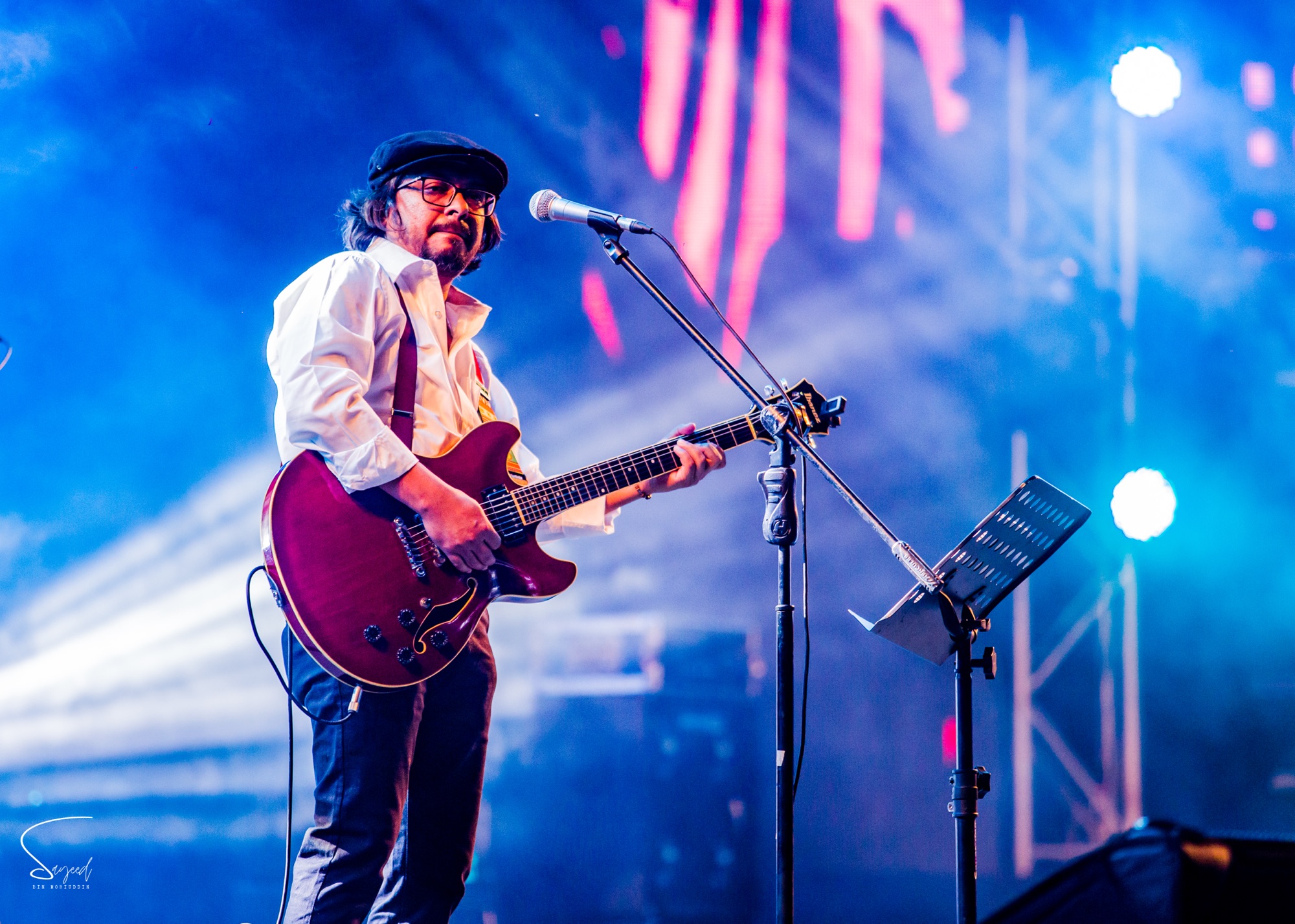 Guitarist in a flat cap playing a red guitar under blue stage light at a live concert in Dhaka — Sayeed BM