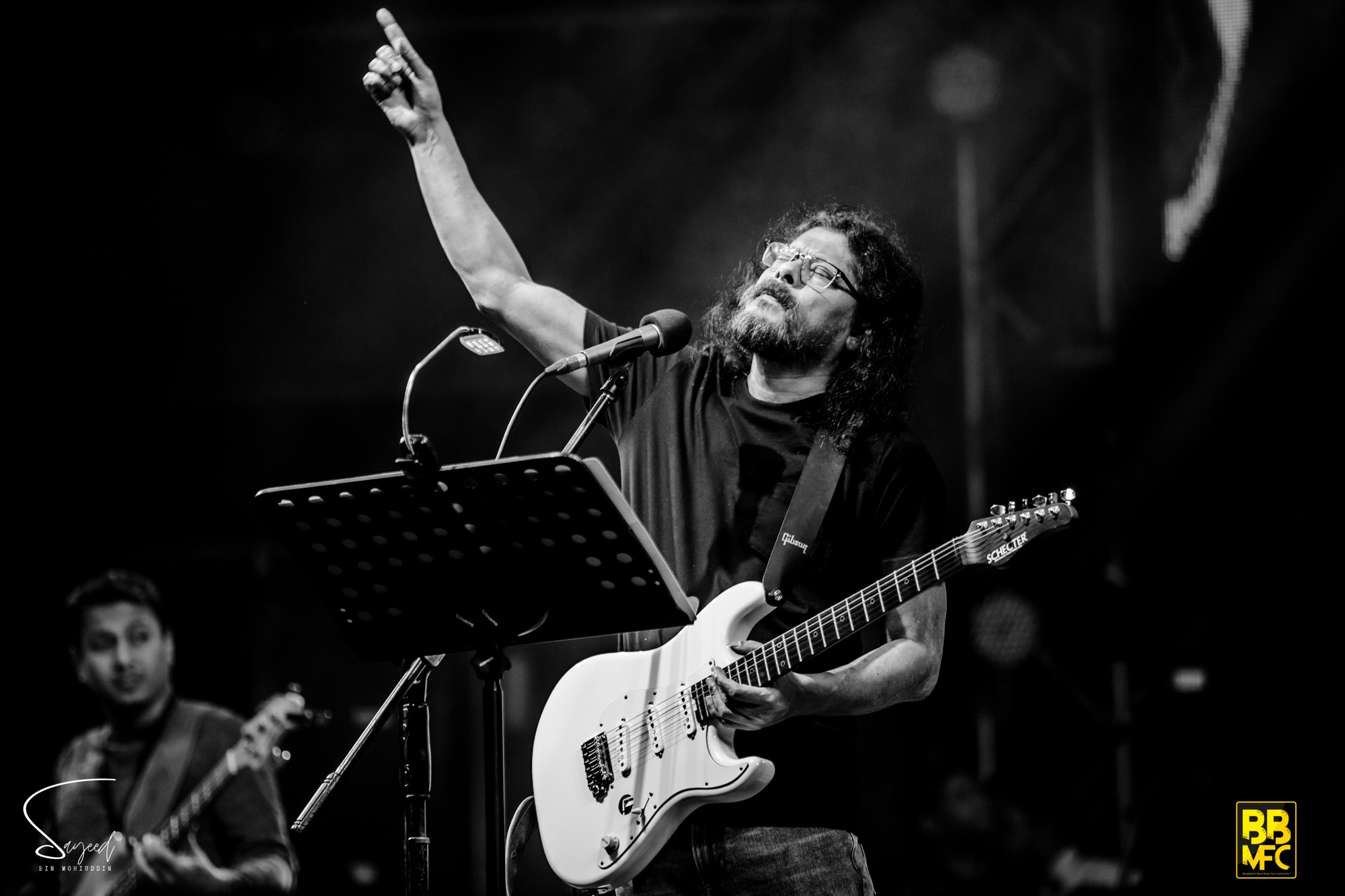 Guitarist raising a fist mid-song with a white Schecter Strat, black-and-white shot from a live concert in Dhaka — Sayeed BM