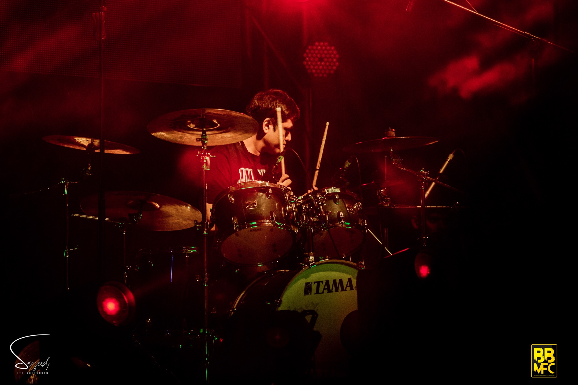 Drummer mid-performance behind a TAMA kit lit in deep red at a live concert in Dhaka — Sayeed BM