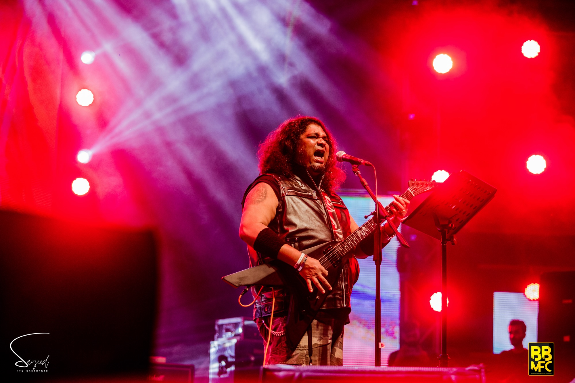 Vocalist belting into the mic with guitar in hand under red and purple light rays at a live concert in Dhaka — Sayeed BM