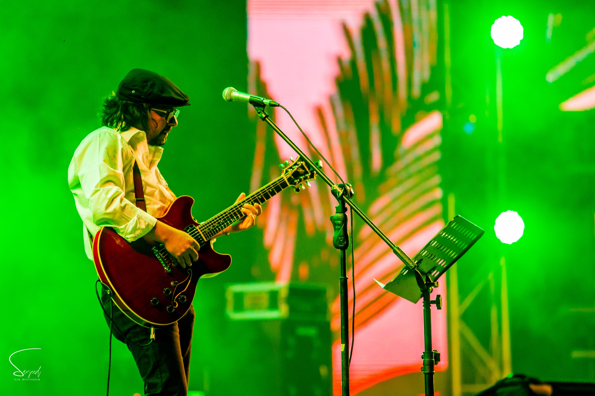Guitarist in a flat cap playing a red semi-hollow guitar under green stage light at a live concert in Dhaka — Sayeed BM