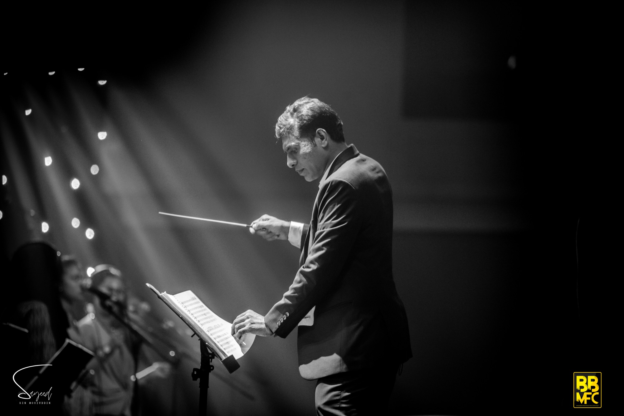 Conductor with baton raised under dramatic light rays, black-and-white, Shironamhin concert in Dhaka — Sayeed BM