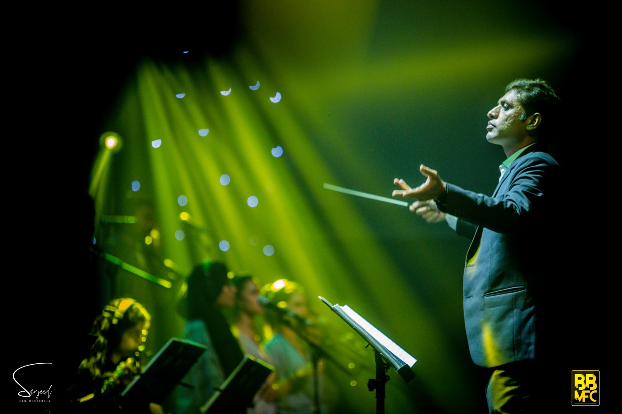 Conductor directing with a baton under green and yellow stage light, choir behind, Shironamhin concert in Dhaka — Sayeed BM