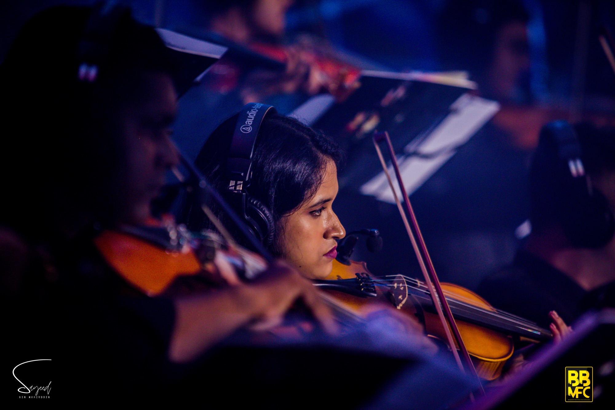 Close-up of a violinist with headphones mid-performance, orange and blue stage light, Shironamhin concert in Dhaka — Sayeed BM