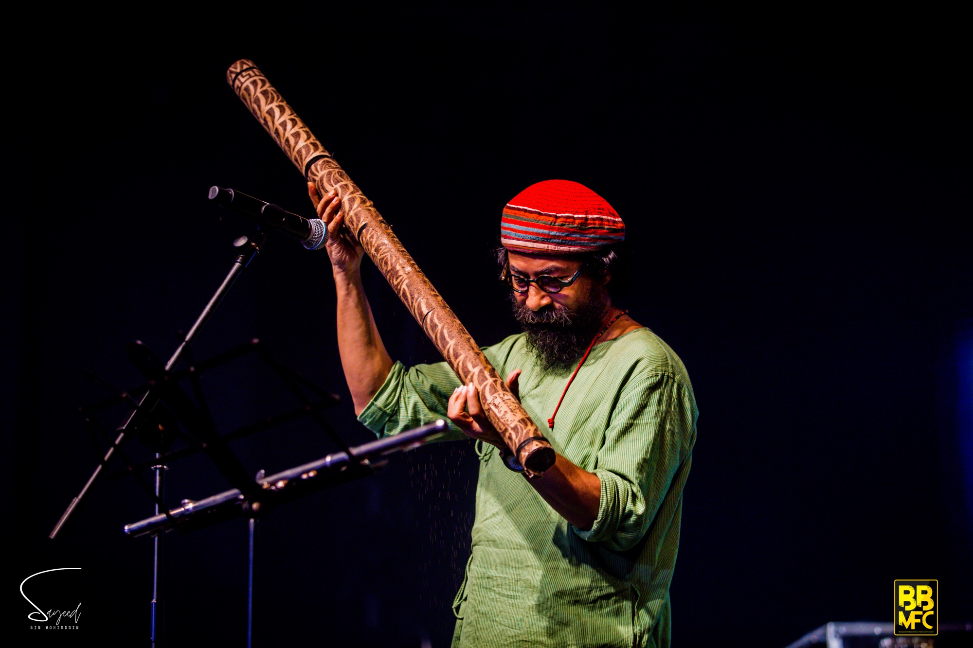 Musician in a red cap playing a didgeridoo against a dark backdrop at a Shironamhin concert in Dhaka — Sayeed BM