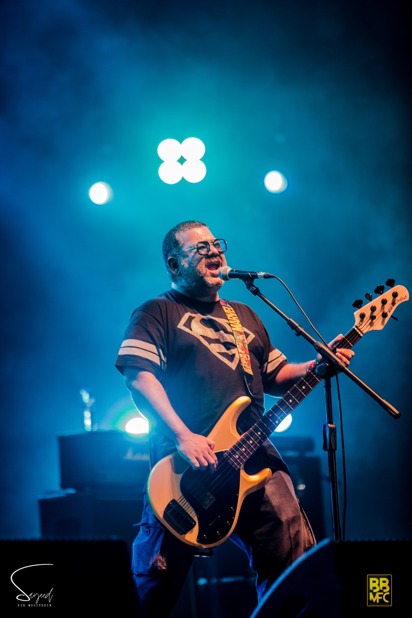 Bassist singing into the mic under teal stage light with a gold bass guitar at a live concert in Dhaka — Sayeed BM